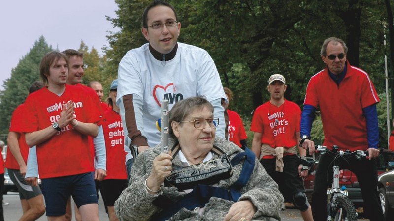 Das Bild zeigt einen Freiwilligendienstleistenden, der eine ältere Frau im Rollstuhl schiebt