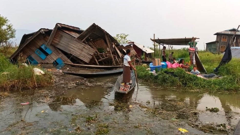 Das Foto zeigt eine Szene nach einer Naturkatastrophe, möglicherweise einem Erdbeben oder einer Überschwemmung. Ein eingestürztes Holzhaus mit blauen Fenstern liegt teilweise im Wasser, während sich Menschen in der Nähe auf einer kleinen erhöhten Fläche mit wenigen Habseligkeiten versammelt haben. Ein Mann steht in einem Boot und scheint sich durch das Wasser zu bewegen. Im Hintergrund sind weitere Holzhäuser auf Stelzen zu sehen, was darauf hindeutet, dass es sich um ein Gebiet mit häufigem Hochwasser handeln könnte. Die Szene vermittelt den Eindruck einer betroffenen Gemeinschaft, die sich nach einer Zerstörung organisiert.