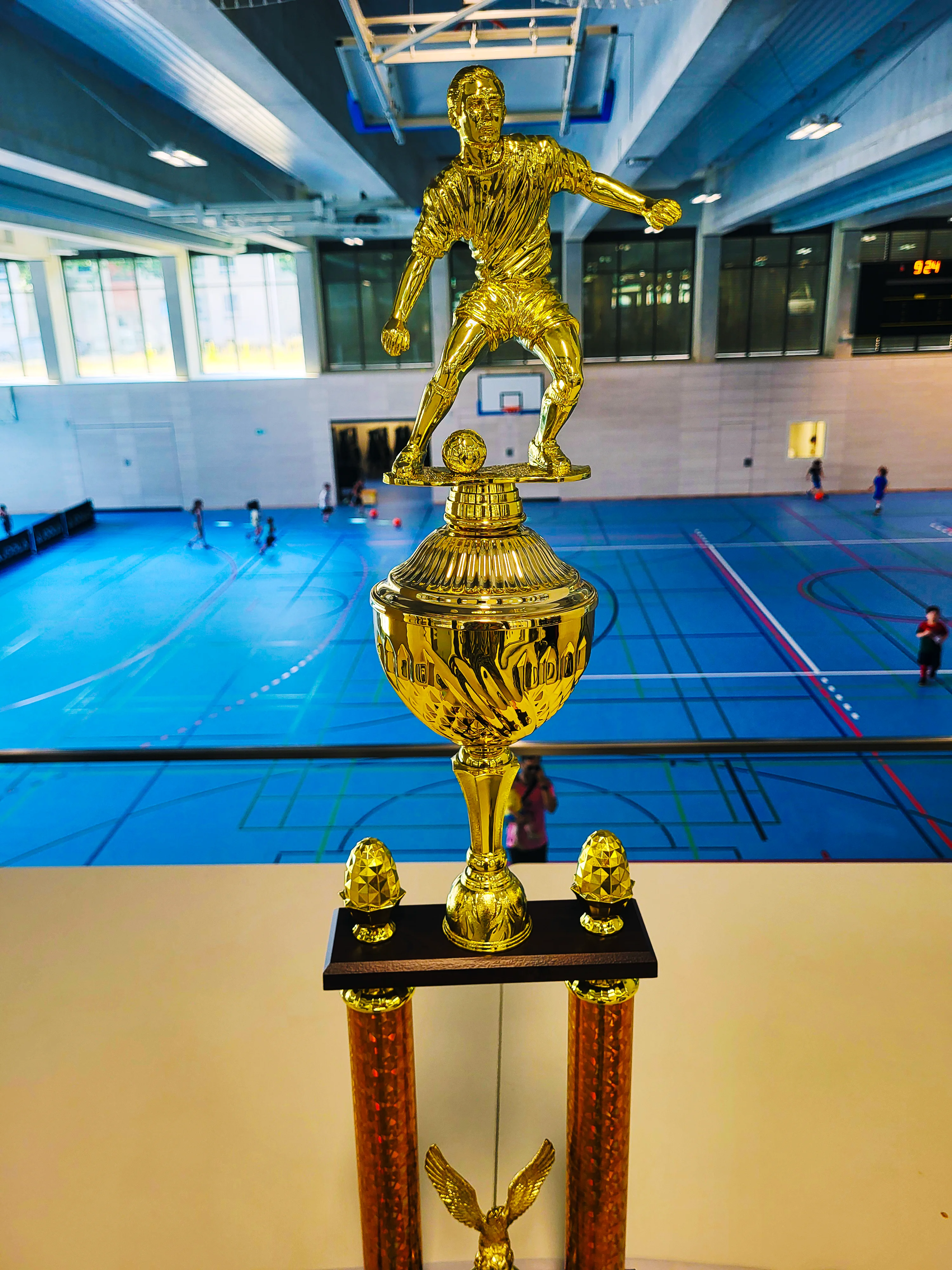 Auf dem Foto ist ein großer, goldfarbener Fußballpokal in einer Sporthalle zu sehen. Im Vordergrund steht der Pokal mit einer Figur eines Fußballspielers auf einem Ball, darunter ein verzierter Kelch und zwei orange-goldene Säulen. Im Hintergrund erkennt man eine blaue Hallenfläche mit eingezeichneten Spielfeldern, Basketballkörben, einigen spielenden Personen sowie große Fensterfronten und die Hallendecke.