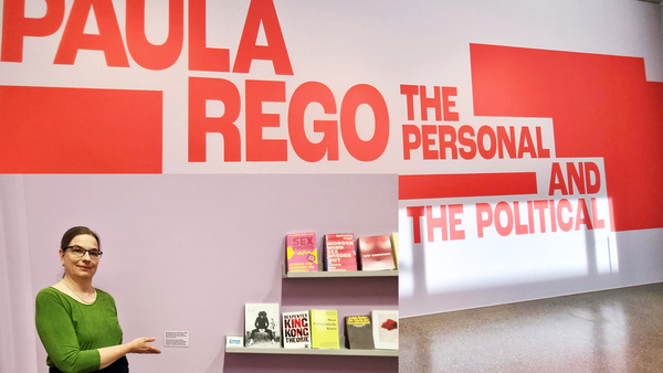 Auf dem Foto ist eine Ausstellung mit dem Titel "Paula Rego: The Personal and the Political" zu sehen. Dieser Titel ist in großen roten Buchstaben auf einer Wand angebracht. Paula Rego war eine bedeutende portugiesisch-britische Künstlerin, bekannt für ihre oft feministisch geprägten Werke. Im Vordergrund steht eine Frau in einem grünen Kleid mit schwarzem Gürtel, die auf ein kleines Wandregal mit Büchern zeigt. Die Bücher scheinen thematisch zur Ausstellung zu passen – viele behandeln feministische oder gesellschaftskritische Themen. Zu erkennen sind unter anderem: „Sex positiv“ „King Kong Theorie“ von Virginie Despentes „Mord ist ihre Leidenschaft“ Weitere Titel zu Gender, Macht und Körperpolitik Die Wand hinter der Frau ist in einem hellen Lila gestrichen, was sich von der weiß-roten Ausstellungsgestaltung abhebt. Es gibt auch ein kleines Schild mit Erklärungstext neben dem Regal, das vermutlich den Kontext der Buchauswahl erläutert.