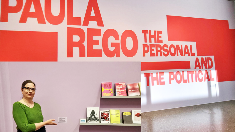 Auf dem Foto ist eine Ausstellung mit dem Titel "Paula Rego: The Personal and the Political" zu sehen. Dieser Titel ist in großen roten Buchstaben auf einer Wand angebracht. Paula Rego war eine bedeutende portugiesisch-britische Künstlerin, bekannt für ihre oft feministisch geprägten Werke. Im Vordergrund steht eine Frau in einem grünen Kleid mit schwarzem Gürtel, die auf ein kleines Wandregal mit Büchern zeigt. Die Bücher scheinen thematisch zur Ausstellung zu passen – viele behandeln feministische oder gesellschaftskritische Themen. Zu erkennen sind unter anderem: „Sex positiv“ „King Kong Theorie“ von Virginie Despentes „Mord ist ihre Leidenschaft“ Weitere Titel zu Gender, Macht und Körperpolitik Die Wand hinter der Frau ist in einem hellen Lila gestrichen, was sich von der weiß-roten Ausstellungsgestaltung abhebt. Es gibt auch ein kleines Schild mit Erklärungstext neben dem Regal, das vermutlich den Kontext der Buchauswahl erläutert.