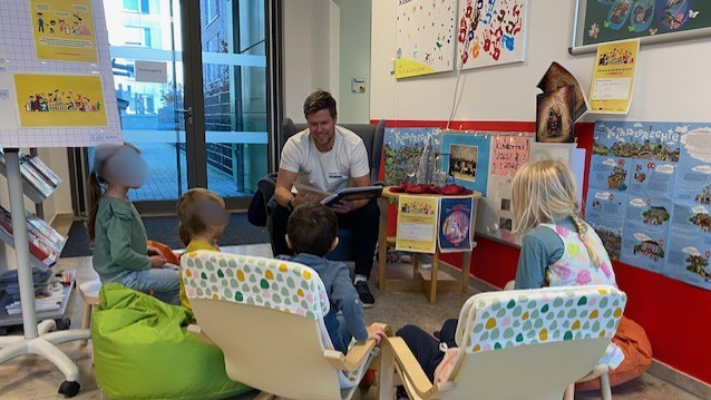 Auf dem Bild ist eine Vorlesesituation in einer Kindertagesstätte (Kita) oder ähnlichen Einrichtung zu sehen. Hier sind die Details: Zentrale Handlung: Ein Erwachsener (vermutlich ein Vater, Erzieher oder Vorlesepate) sitzt in der Mitte und liest aus einem Buch vor, das er in den Händen hält. Er lächelt dabei. Zuhörer: Vier kleine Kinder sitzen um den Vorleser herum und hören zu. Sitzmöglichkeiten: Die Kinder sitzen bequem in verschiedenen Stühlen/Sitzgelegenheiten: Links: Ein Kind sitzt auf einem grünen Sitzsack. Mitte und Rechts: Drei Kinder sitzen in niedrigen, holzgerahmten Kindersesseln mit einem hellen, gemusterten Stoffbezug. Hintergrund und Dekoration: Im Hintergrund sind Poster an der Wand zu sehen, die auf eine Kindereinrichtung hindeuten. Eines der Poster fragt: "Kinder haben Rechte!" und zeigt bunte Handabdrücke. Auf einem kleinen Tisch hinter dem Vorleser stehen weitere Bücher und Dekorationsgegenstände. Ganz links steht ein Flipchart oder Plakatständer mit Grafiken, die ebenfalls kinderbezogene Themen (vermutlich Kinderrechte oder Regeln) behandeln. Im linken Hintergrund ist eine Glastür zu sehen, durch die Tageslicht einfällt und ein Außenbereich erkennbar ist.