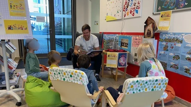 Auf dem Bild ist eine Vorlesesituation in einer Kindertagesstätte (Kita) oder ähnlichen Einrichtung zu sehen. Hier sind die Details: Zentrale Handlung: Ein Erwachsener (vermutlich ein Vater, Erzieher oder Vorlesepate) sitzt in der Mitte und liest aus einem Buch vor, das er in den Händen hält. Er lächelt dabei. Zuhörer: Vier kleine Kinder sitzen um den Vorleser herum und hören zu. Sitzmöglichkeiten: Die Kinder sitzen bequem in verschiedenen Stühlen/Sitzgelegenheiten: Links: Ein Kind sitzt auf einem grünen Sitzsack. Mitte und Rechts: Drei Kinder sitzen in niedrigen, holzgerahmten Kindersesseln mit einem hellen, gemusterten Stoffbezug. Hintergrund und Dekoration: Im Hintergrund sind Poster an der Wand zu sehen, die auf eine Kindereinrichtung hindeuten. Eines der Poster fragt: "Kinder haben Rechte!" und zeigt bunte Handabdrücke. Auf einem kleinen Tisch hinter dem Vorleser stehen weitere Bücher und Dekorationsgegenstände. Ganz links steht ein Flipchart oder Plakatständer mit Grafiken, die ebenfalls kinderbezogene Themen (vermutlich Kinderrechte oder Regeln) behandeln. Im linken Hintergrund ist eine Glastür zu sehen, durch die Tageslicht einfällt und ein Außenbereich erkennbar ist.