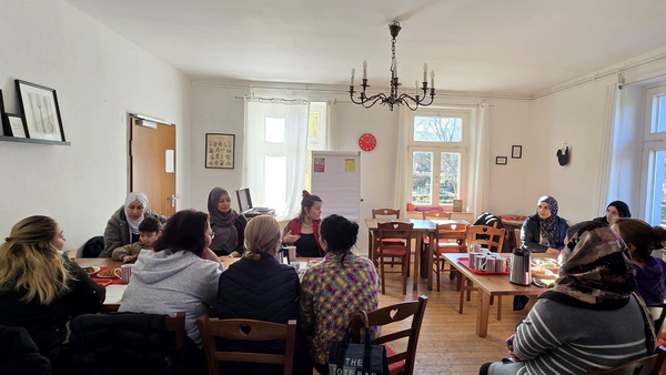 Auf dem Bild ist eine Gruppe von Menschen zu sehen, die gemeinsam in einem hellen Raum an einem großen Tisch sitzen und sich unterhalten. Es wirkt wie ein Treffen oder Kurs in gemütlicher Café- oder Gruppenraum-Atmosphäre mit Kaffee, Snacks und Flipchart im Hintergrund.