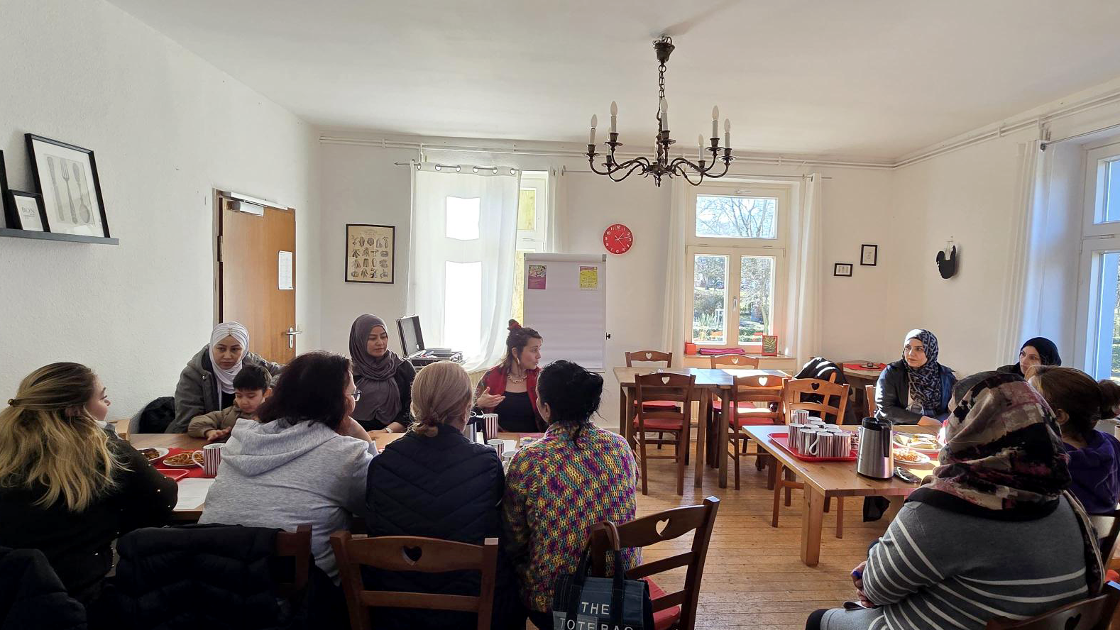 Auf dem Bild ist eine Gruppe von Menschen zu sehen, die gemeinsam in einem hellen Raum an einem großen Tisch sitzen und sich unterhalten. Es wirkt wie ein Treffen oder Kurs in gemütlicher Café- oder Gruppenraum-Atmosphäre mit Kaffee, Snacks und Flipchart im Hintergrund.