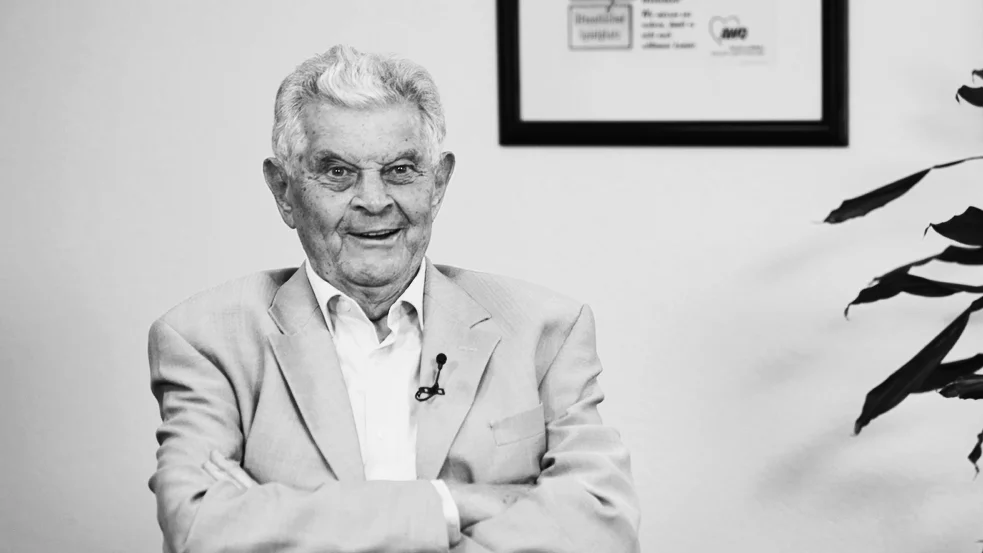 Auf dem schwarz-weiß Foto ist Paul Saatkamp zu sehen, der mit verschränkten Armen sitzt. Er trägt einen hellen Anzug. Im Hintergrund sind an der Wand gerahmte Zertifikate und ein Teil einer Pflanze zu sehen.