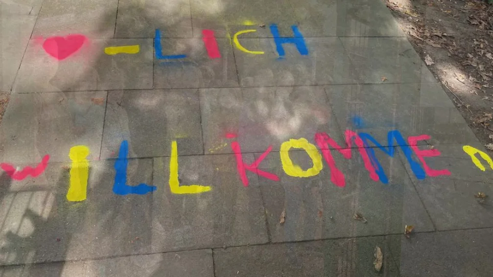 Das Foto zeigt einen Gehweg mit in verschiedenen Farben aufgemalten Worten. Der Text lautet “-[Herzsymbol]LICH WLLKOMMEN,”