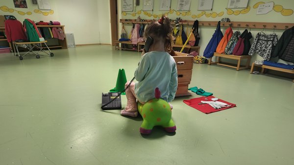Auf dem Bild sitzt ein Kind mit dem Rücken zur Kamera auf einem grünen Hüpftier und hält den Hörer eines schwarzen Tischtelefons ans Ohr. Das Telefon steht daneben auf dem Boden; eine Spiralschnur verbindet den Hörer mit dem Gerät. Die Szene spielt in einem Garderobenbereich: An den Wänden hängen Jacken, Taschen und Rucksäcke über niedrigen Holzbänken. Im Raum stehen außerdem eine Holzkiste und verschiedene Spielmaterialien. Das Gesicht des Kindes ist nicht zu erkennen.