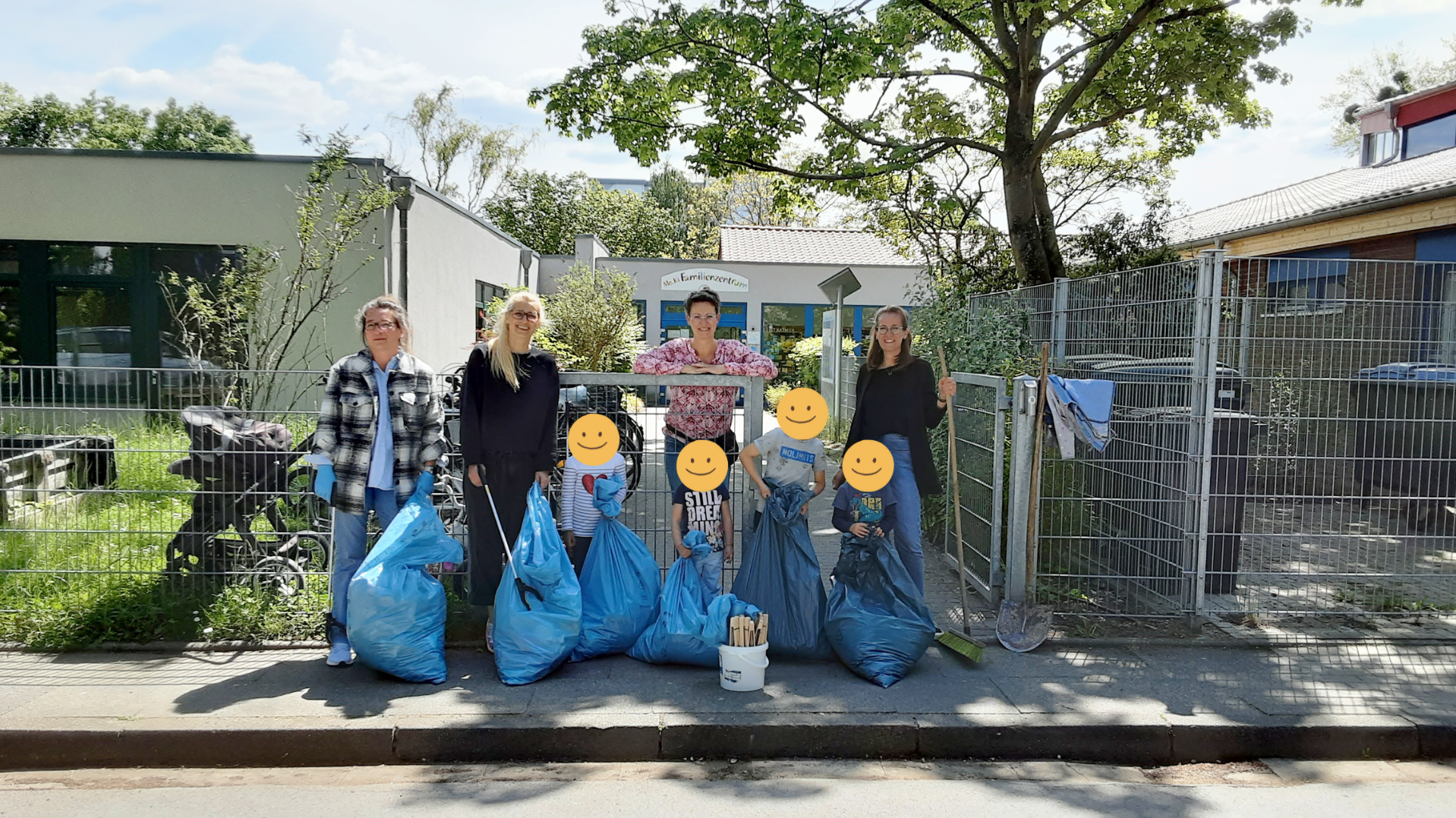 Auf dem Foto sind fünf Erwachsene und drei Kinder zu sehen, die anscheinend Müll gesammelt haben. Sie stehen vor einem Zaun mit mehreren blauen Müllsäcken, einem Eimer mit Greifzangen und Besen. Im Hintergrund sind Gebäude und Bäume zu erkennen. Die Gesichter der Kinder sind mit gelben Smiley-Aufklebern verdeckt.