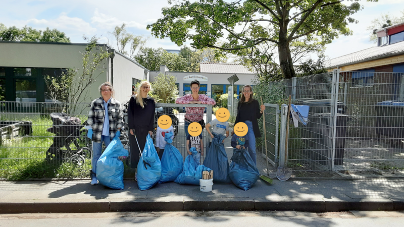 Auf dem Foto sind fünf Erwachsene und drei Kinder zu sehen, die anscheinend Müll gesammelt haben. Sie stehen vor einem Zaun mit mehreren blauen Müllsäcken, einem Eimer mit Greifzangen und Besen. Im Hintergrund sind Gebäude und Bäume zu erkennen. Die Gesichter der Kinder sind mit gelben Smiley-Aufklebern verdeckt.