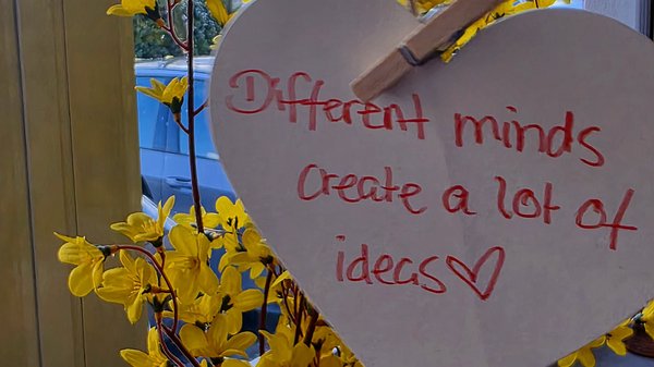 Auf dem Bild sieht man gelbe Blütenzweige vor einem Fenster, daran hängt ein Herz aus Pappe mit der handschriftlichen Aufschrift „Different minds create a lot of ideas“ und einem kleinen Herzsymbol.