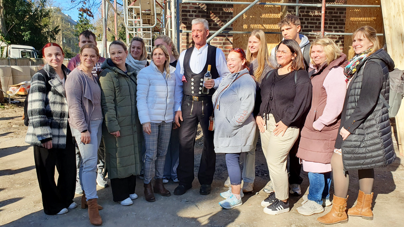 Auf dem Bild steht eine Gruppe von Menschen gemeinsam auf einer Baustelle vor einem im Bau befindlichen Gebäude. In der Mitte steht ein Mann in traditioneller Zimmerer- bzw. Handwerkerkleidung und hält eine Flasche in der Hand; um ihn herum gruppieren sich mehrere Personen in Alltagskleidung, die offenbar zu einem gemeinsamen Anlass wie etwa einem Richtfest zusammengekommen sind.