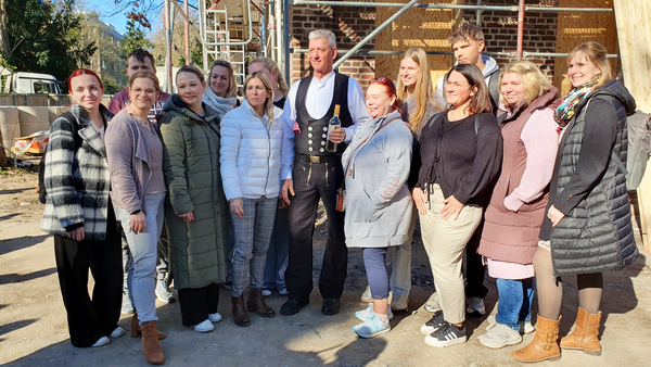Auf dem Bild steht eine Gruppe von Menschen gemeinsam auf einer Baustelle vor einem im Bau befindlichen Gebäude. In der Mitte steht ein Mann in traditioneller Zimmerer- bzw. Handwerkerkleidung und hält eine Flasche in der Hand; um ihn herum gruppieren sich mehrere Personen in Alltagskleidung, die offenbar zu einem gemeinsamen Anlass wie etwa einem Richtfest zusammengekommen sind.