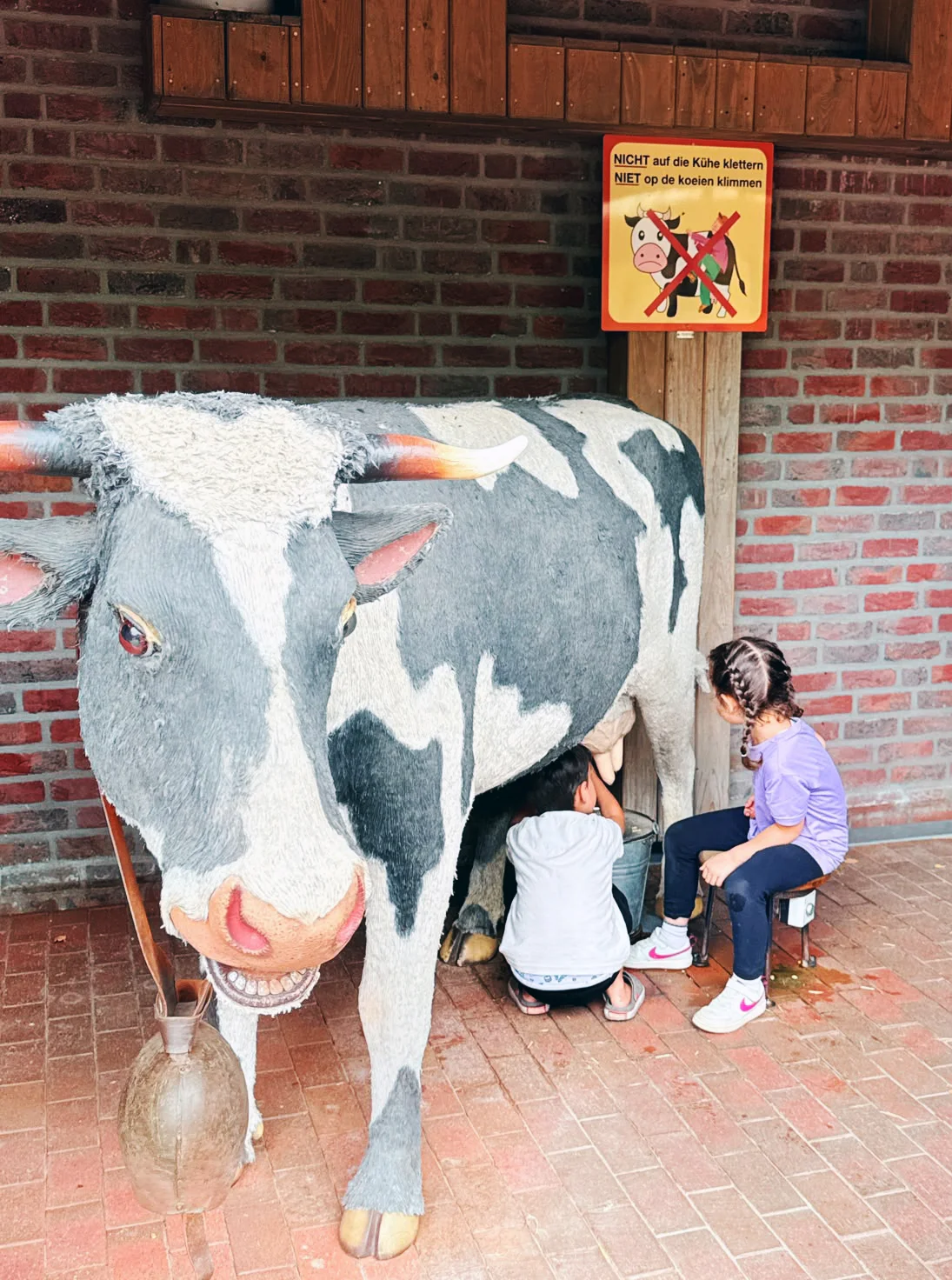 Auf dem Bild ist eine lebensgroße, schwarz-weiß gefleckte Kuh-Figur in einem stallähnlichen Raum mit roter Backsteinwand zu sehen. Zwei Kinder sitzen bzw. hocken daneben und spielen offenbar das Melken der Kuh mit einem darunterstehenden Eimer nach. Über der Figur hängt ein gelbes Warnschild auf Deutsch und Niederländisch: „Nicht auf die Kühe klettern / Niet op de koeien klimmen“
