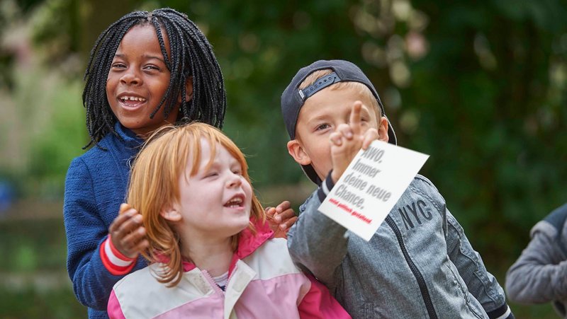 Drei spielende Kinder. Ein Junge hält einen Zettel in der Hand mit dem Schriftzug "AWO. Immer Deine neue Chance"