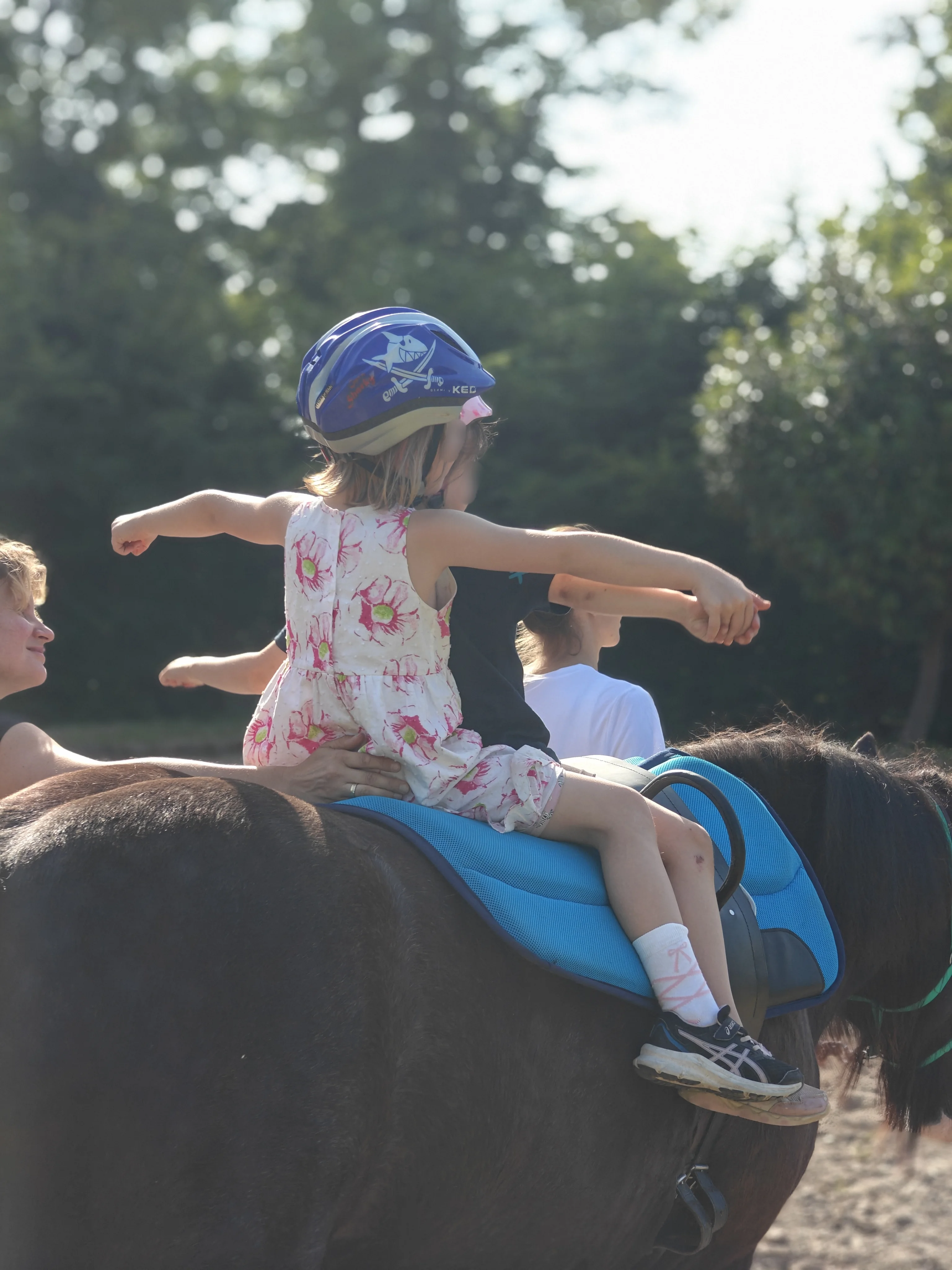 Ein Kind sitzt mit Helm und Sommerkleid auf einem dunklen Pony. Erwachsene sichern das Kind seitlich, während es mit ausgestreckten Armen eine Übung auf dem Pony macht (Balance-/Vertrauensübung). Die Szene spielt draußen auf einem Reitplatz bei sonnigem Wetter.