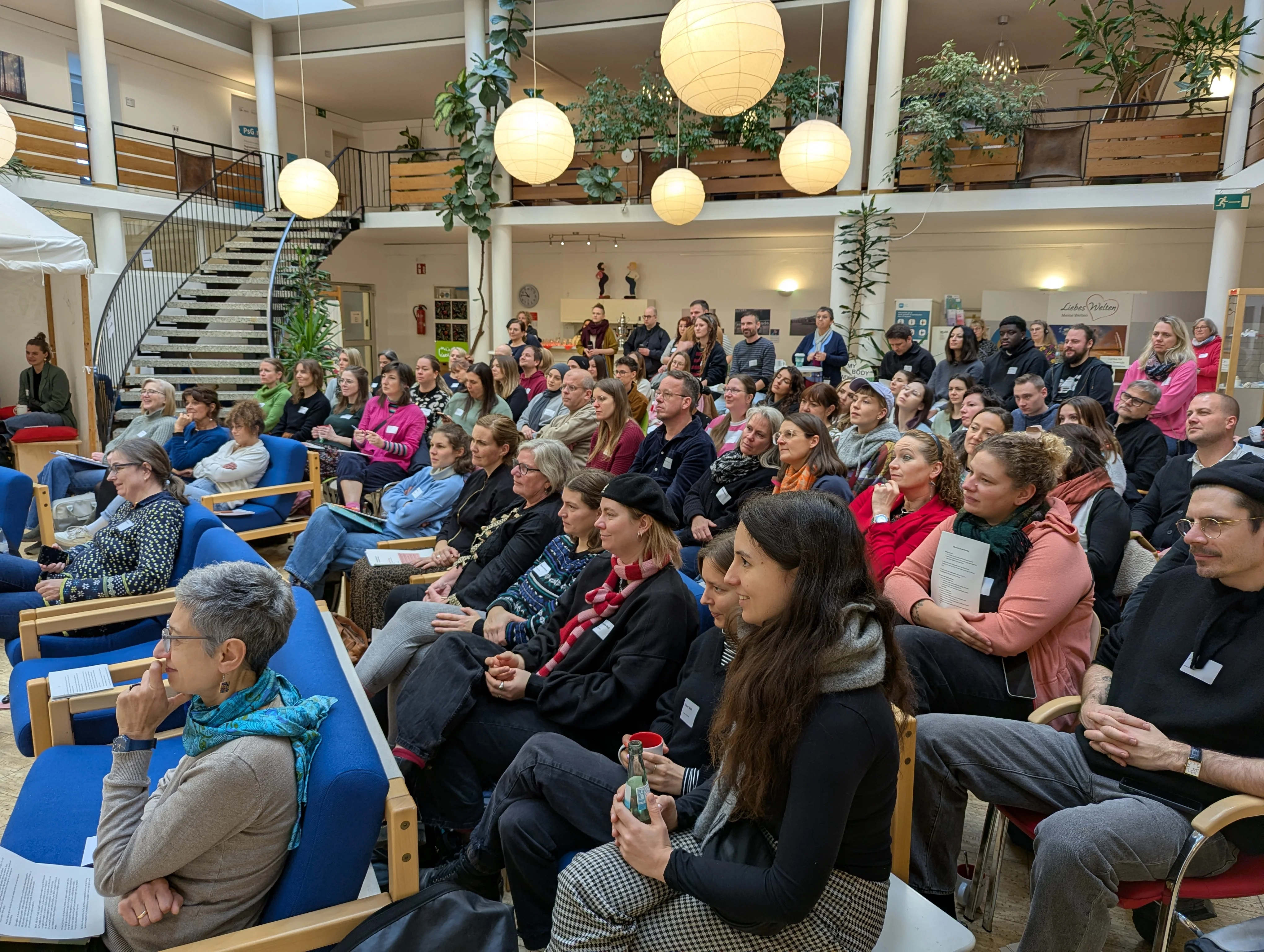 Auf dem Foto ist eine Gruppe von Menschen zu sehen, die an einer Veranstaltung oder Konferenz teilnehmen. Hier sind die Details: Publikum: Der Hauptfokus liegt auf dem Publikum, das in Stuhlreihen sitzt und aufmerksam in eine Richtung blickt, was darauf hindeutet, dass sie einem Vortrag oder einer Präsentation folgen (die auf diesem Bild nicht direkt sichtbar ist). Veranstaltungsort: Die Veranstaltung findet in einem großen, hellen Innenhof oder Atrium eines Gebäudes statt. Das Gebäude hat mindestens zwei Stockwerke, mit umlaufenden Balkonen oder Galerien. Eine markante schwarze Wendeltreppe ist auf der linken Seite sichtbar. Die Umgebung ist mit vielen Pflanzen dekoriert, was eine angenehme Atmosphäre schafft. Über den Köpfen der Zuhörer hängen mehrere große, runde, leuchtende Papier- oder Stofflaternen (Lampions). Atmosphäre: Die Kleidung und Haltung der Menschen deutet auf eine geschäftliche oder bildungsbezogene Veranstaltung hin (wie ein Fachtag, passend zum vorherigen Bild). Einige halten Notizen oder Broschüren. Es handelt sich um eine Innenaufnahme des Publikums während einer Veranstaltung in einem modernen, begrünten Atrium.