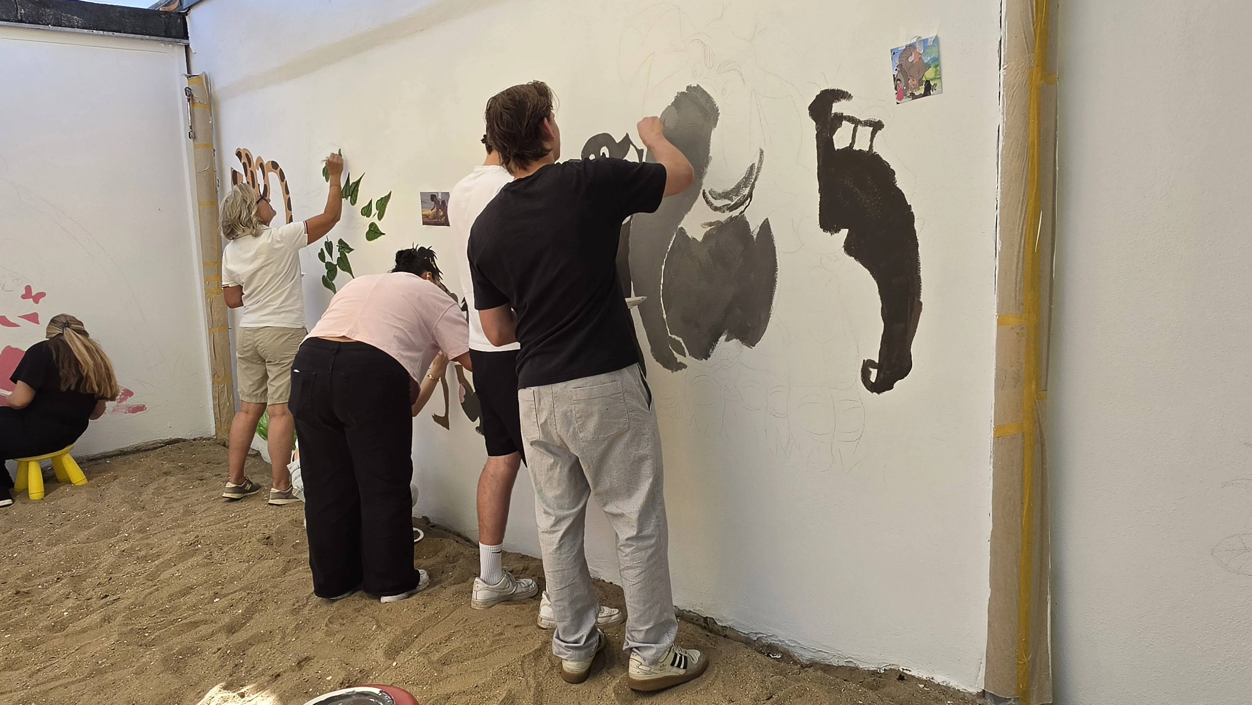 Auf dem Bild sind mehrere Personen zu sehen, die eine weiße Wand in einem sandigen Außenbereich bemalen. Links sitzen und stehen einige Menschen vor bereits vorgezeichneten bzw. leicht kolorierten Motiven, weiter rechts malen zwei Personen an großen, dunklen Tierfiguren und grünen Blättern. Man erkennt, dass auf der Wand Konturen von Comic‑/Tierfiguren (unter anderem ein Elefant und eine dunkle Gestalt) skizziert sind, die gerade ausgemalt werden; an der Wand hängt zur Orientierung ein kleines Referenzbild.