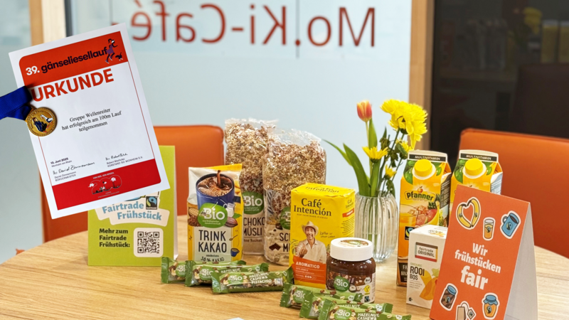 Auf dem Foto ist ein liebevoll arrangierter Frühstückstisch zu sehen, der ganz im Zeichen von Fairtrade steht. Zu erkennen sind: Fairtrade-Produkte, darunter: dmBio Trinkkakao Choco- und Schokomüsli Café Intención Kaffee Rooibos-Tee von Fairtrade Original Bio Haselnuss-Cashew-Pistazien-Riegel von dmBio Bio Nuss-Nougat-Creme Pfanner Multivitaminsaft Dekoration: Ein frischer Blumenstrauß mit gelben und roten Blüten in einer Glasvase Ein Aufsteller mit der Aufschrift „Wir frühstücken fair“ Ein weiterer Aufsteller mit QR-Code und dem Hinweis auf das Fairtrade Frühstück Im Vordergrund links ist eine Urkunde zu sehen, mit einer Medaille: 39. Gänseliesellauf Ausgezeichnet wurde die Gruppe Wellenreiter für ihre erfolgreiche Teilnahme am 100m-Lauf am 13. Juni 2025 Im Hintergrund erkennt man durch die Glasscheibe die Aufschrift „Mo.Ki-Café“, was auf den Veranstaltungsort hinweist