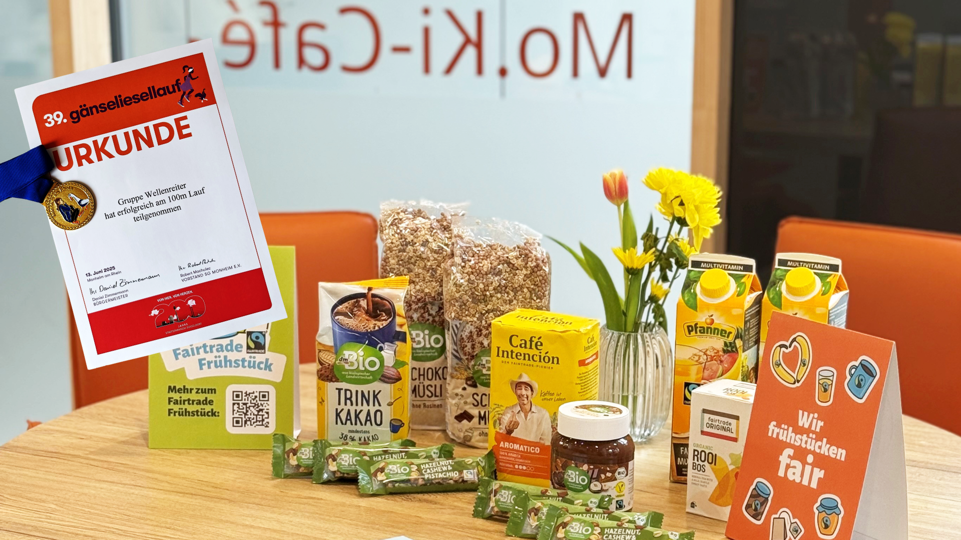 Auf dem Foto ist ein liebevoll arrangierter Frühstückstisch zu sehen, der ganz im Zeichen von Fairtrade steht. Zu erkennen sind: Fairtrade-Produkte, darunter: dmBio Trinkkakao Choco- und Schokomüsli Café Intención Kaffee Rooibos-Tee von Fairtrade Original Bio Haselnuss-Cashew-Pistazien-Riegel von dmBio Bio Nuss-Nougat-Creme Pfanner Multivitaminsaft Dekoration: Ein frischer Blumenstrauß mit gelben und roten Blüten in einer Glasvase Ein Aufsteller mit der Aufschrift „Wir frühstücken fair“ Ein weiterer Aufsteller mit QR-Code und dem Hinweis auf das Fairtrade Frühstück Im Vordergrund links ist eine Urkunde zu sehen, mit einer Medaille: 39. Gänseliesellauf Ausgezeichnet wurde die Gruppe Wellenreiter für ihre erfolgreiche Teilnahme am 100m-Lauf am 13. Juni 2025 Im Hintergrund erkennt man durch die Glasscheibe die Aufschrift „Mo.Ki-Café“, was auf den Veranstaltungsort hinweist 