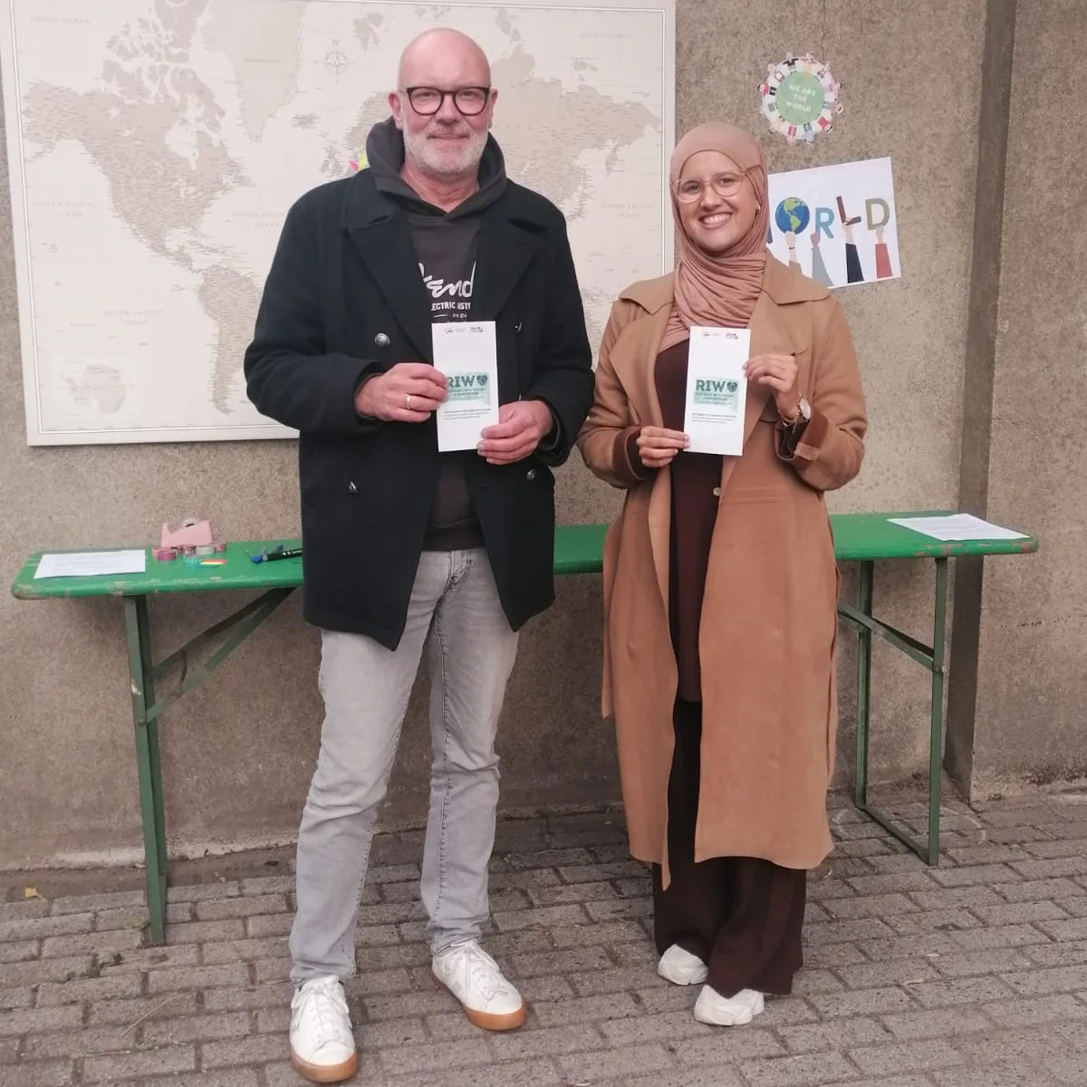 Auf dem Foto sind zwei Personen zu sehen, die nebeneinander vor einer grauen Wand stehen und Broschüren oder Faltblätter in die Kamera halten. Die linke Person ist ein Mann mit Brille, Glatze und Bart. Er trägt einen schwarzen Mantel über einem Kapuzenpullover, hellgraue Jeans und weiße Sportschuhe. Die rechte Person ist eine Frau, die ein hellbraunes Kopftuch (Hijab) und einen langen, beigen oder hellbraunen Mantel über brauner Kleidung trägt. Sie trägt ebenfalls weiße Schuhe. Beide Personen halten ein weißes Faltblatt mit dem Logo "RIW" in den Händen, ähnlich der Broschüre auf dem vorherigen Foto. Im Hintergrund ist Folgendes zu erkennen: Eine große Weltkarte hängt an der Wand. Ein grüner Klapptisch steht vor der Wand. Rechts von der Karte ist eine Dekoration mit dem Wort "WORLD" und weiteren farbigen Elementen an der Wand befestigt. Der Boden besteht aus Pflastersteinen.