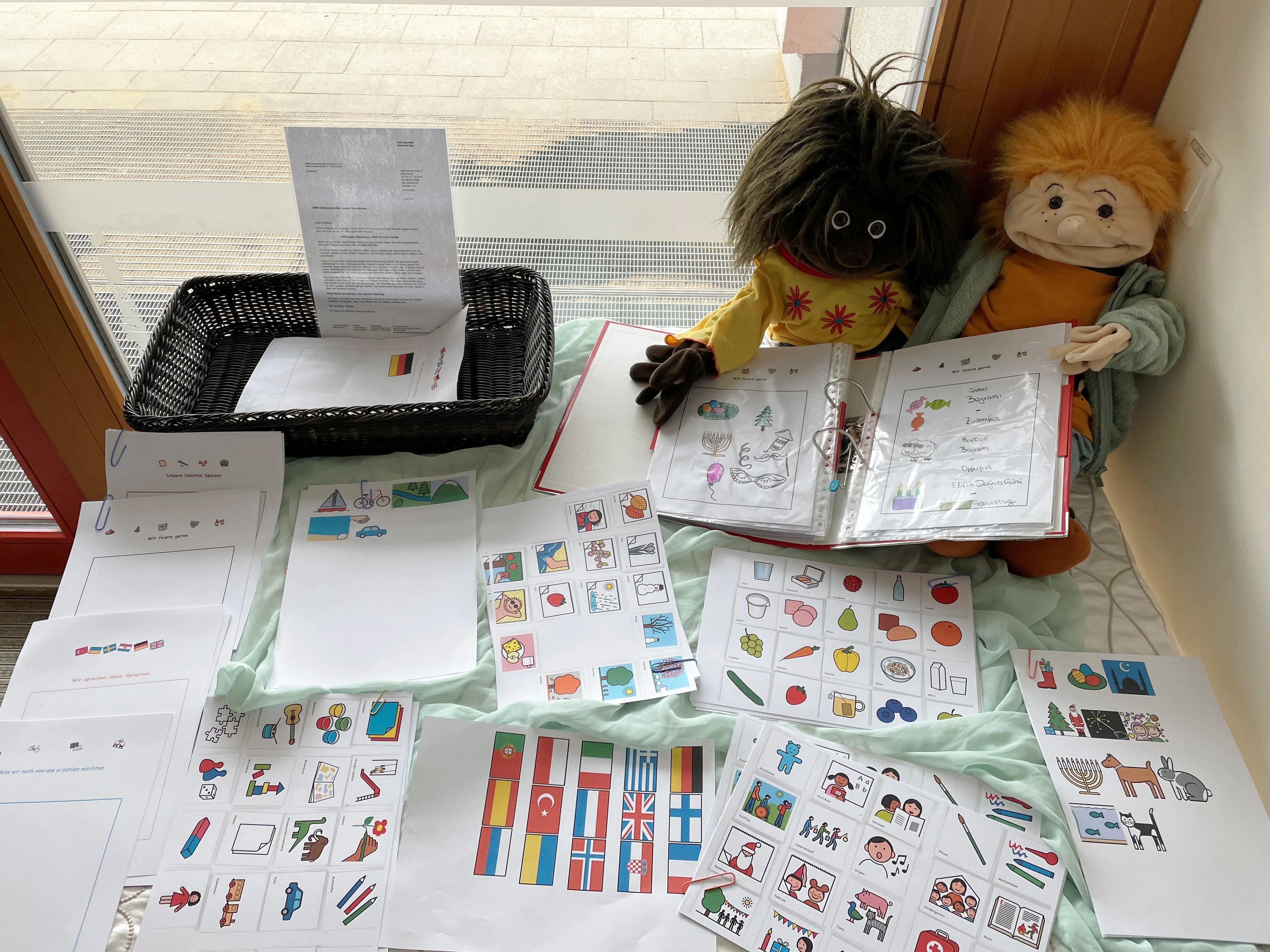 Auf dem Bild ist ein pädagogisches Arbeitsmaterial-Set zu sehen, das auf einem Tisch ausgebreitet ist. Im Einzelnen sind folgende Elemente erkennbar: Zwei Handpuppen im Hintergrund: Eine mit dunklem Haar und brauner Haut (in einem bunten gelben Oberteil) und eine mit orangefarbenem Haar – typische Kita-Handpuppen zur spielerischen Kommunikation mit Kindern. Ein aufgeklapptes Ringbuch/Ordner, den eine der Puppen hält – darin befinden sich Bildkarten mit Symbolen, z. B. zu Festen und Feiertagen (erkennbar sind u. a. Luftballons, Süßigkeiten, Kerzen sowie handgeschriebene Begriffe wie „Zuckerfest", „Karneval/Bajram", „Chanukka"). Zahlreiche ausgebreitete Arbeitsblätter mit piktogrammartigen Bildkarten zu verschiedenen Themen, darunter: Lebensmittel und Alltagsgegenstände Familienkonstellationen und Menschen Freizeitaktivitäten und Spielzeug Religiöse und kulturelle Symbole (z. B. Weihnachtsbaum, Menora, Halbmond) Ein Blatt mit internationalen Flaggen (u. a. Portugal, Türkei, Italien, Griechenland, Großbritannien, Finnland, Norwegen, Kroatien) – als Hinweis auf Mehrsprachigkeit und Interkulturalität. Ein schwarzer Korb mit Briefbögen (vermutlich Elternbriefe) sowie ein Dokument mit der deutschen Flagge, das ein Anschreiben zu sein scheint.