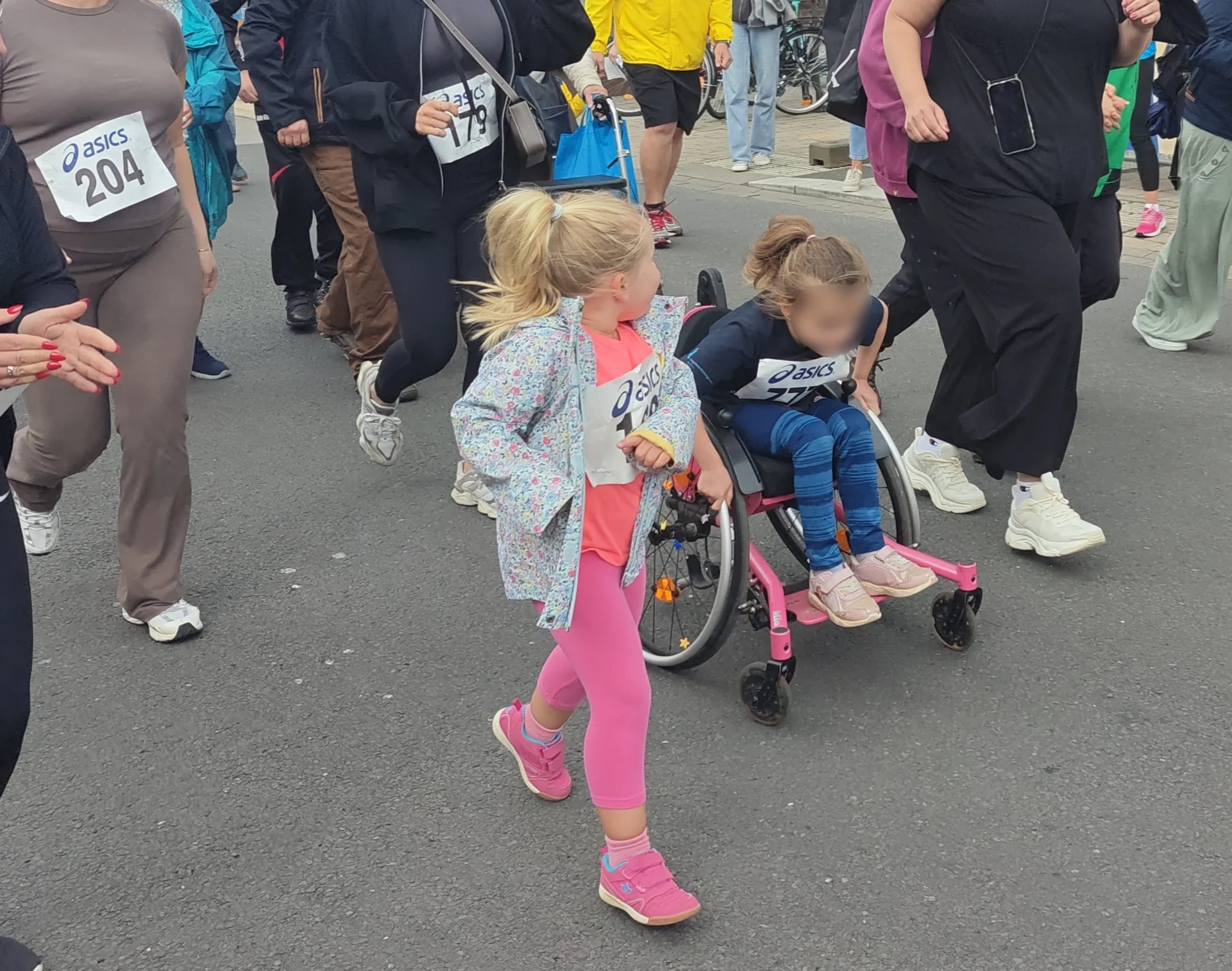 Auf dem Bild ist eine inklusive Laufszene bei einer Laufveranstaltung zu sehen. Im Mittelpunkt schiebt ein Mädchen im pinken Sportoutfit ein anderes Mädchen im Rollstuhl, beide tragen Startnummern eines Laufes. Rundherum laufen weitere Erwachsene und Kinder mit Startnummern auf der abgesperrten Straße mit, was den Charakter eines gemeinsamen Familien- oder Sponsorenlaufs unterstreicht.