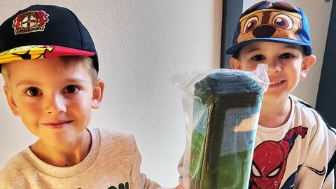 Zu sehen sind zwei lächelnde Jungen in einer Wohnung. Der Junge links trägt ein Pokémon-Shirt und eine schwarze Kappe mit rotem Schirm. Der Junge rechts trägt ein Spiderman-Shirt sowie eine Kappe mit einem Comic-Motiv. Gemeinsam halten sie einen zusammengerollten, noch in Folie verpackten Teppich oder Läufer hoch.