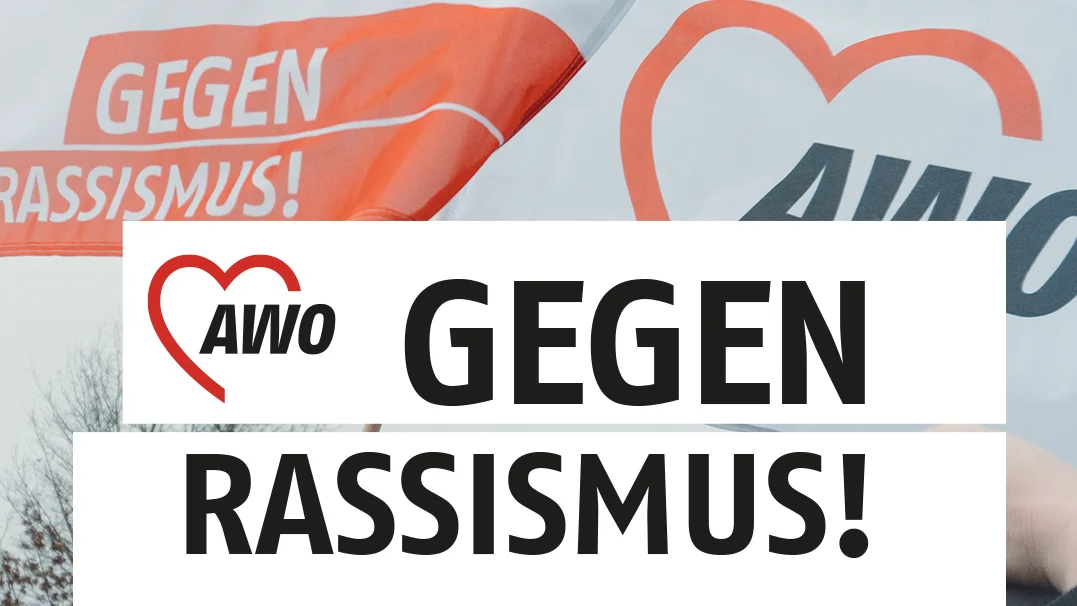Auf dem Foto sind zwei Fahnen der Arbeiterwohlfahrt (AWO) zu sehen, die gegen Rassismus werben. Die Fahnen sind weiß mit einem orangefarbenen Herz, das das AWO-Logo enthält. Unter dem Logo steht in schwarzen Buchstaben „GEGEN RASSISMUS!“. Im Vordergrund des Fotos sind die Hände von Menschen zu sehen, die die Fahnen hochhalten. Die Menschen sind nicht identifizierbar. Im Hintergrund sind weitere Menschen zu sehen, die offenbar an einer Demonstration oder Kundgebung teilnehmen. Das Foto ist bei Tageslicht aufgenommen und hat eine neutrale Farbgebung. Die Stimmung des Fotos ist positiv und kämpferisch.