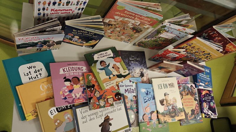 Auf dem Bild ist eine Sammlung von Kinderbüchern zu sehen, die auf einem grünen Tisch ausgebreitet sind. Die Bücher sind auf Deutsch und behandeln verschiedene Themen, die für Kinder relevant sind: Diversität und Inklusion: Bücher wie "Kleidung ist für alle da" und "Charlie und der Schokoladenhund" Identität: "Ich bin Mara" Spiel und Alltag: "Komm, wir uns!", "Hier kommt Mila!", "Kannst du wirsagen" Fantasie und Geschichten: "Das Herz von Elfland", verschiedene illustrierte Kinderbücher Die Bücher haben bunte, kindgerechte Cover mit vielfältigen Illustrationen von Kindern unterschiedlicher Herkunft. Es scheint sich um eine bewusst zusammengestellte Sammlung zu handeln, die Themen wie Vielfalt, Gleichberechtigung und Selbstfindung behandelt - passend für eine Bildungs- oder Sozialeinrichtung