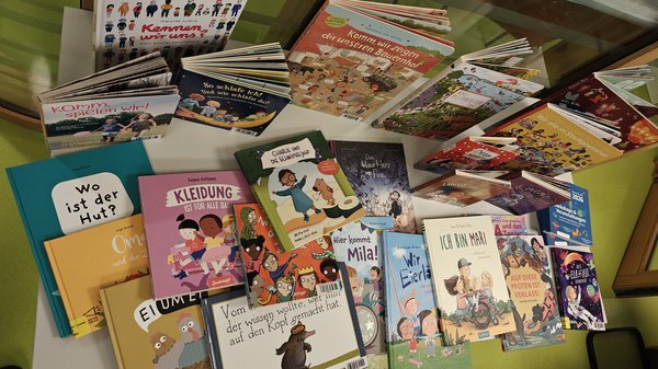 Auf dem Bild ist eine Sammlung von Kinderbüchern zu sehen, die auf einem grünen Tisch ausgebreitet sind. Die Bücher sind auf Deutsch und behandeln verschiedene Themen, die für Kinder relevant sind: Diversität und Inklusion: Bücher wie "Kleidung ist für alle da" und "Charlie und der Schokoladenhund" Identität: "Ich bin Mara" Spiel und Alltag: "Komm, wir uns!", "Hier kommt Mila!", "Kannst du wirsagen" Fantasie und Geschichten: "Das Herz von Elfland", verschiedene illustrierte Kinderbücher Die Bücher haben bunte, kindgerechte Cover mit vielfältigen Illustrationen von Kindern unterschiedlicher Herkunft. Es scheint sich um eine bewusst zusammengestellte Sammlung zu handeln, die Themen wie Vielfalt, Gleichberechtigung und Selbstfindung behandelt - passend für eine Bildungs- oder Sozialeinrichtung