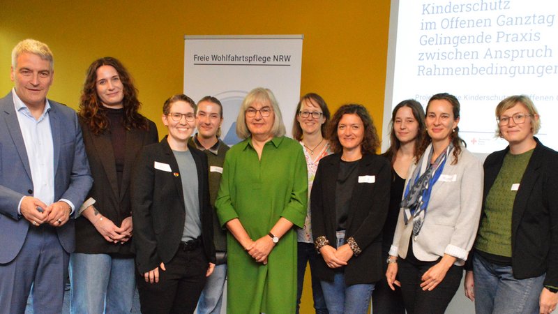 Auf dem Bild ist eine Gruppenaufnahme mit zehn Personen zu sehen. Sie stehen in einem Veranstaltungsraum nebeneinander und schauen in die Kamera. Die Person in der Mitte trägt ein auffälliges grünes Kleid. Im Hintergrund stehen ein Roll-up mit der Aufschrift „Freie Wohlfahrtspflege NRW“ und eine Projektionsfläche. Auf dieser sind unter anderem „Kinderschutz im Offenen Ganztag“ sowie „Gelingende Praxis“ und „Rahmenbedingungen“ zu lesen. Das Foto wirkt daher wie eine Gruppenaufnahme im Rahmen einer Fachveranstaltung zum Kinderschutz im offenen Ganztag. Mehrere Personen tragen Namensschilder; ihre Namen sind auf dem Bild jedoch nicht zuverlässig lesbar.