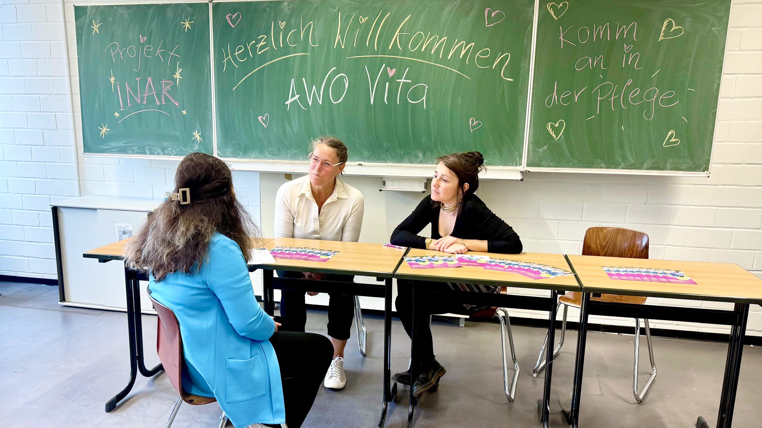  Auf dem Foto sind drei Personen an einem Tisch in einem Klassenraum zu sehen. An der grünen Tafel hinter ihnen stehen verschiedene Sätze, wie z. B. „Projekt INAR“, „Herzlich Willkommen AWO Vita“ und „Komm an in der Pflege“. Auf den Tischen liegen Informationsmaterialien.