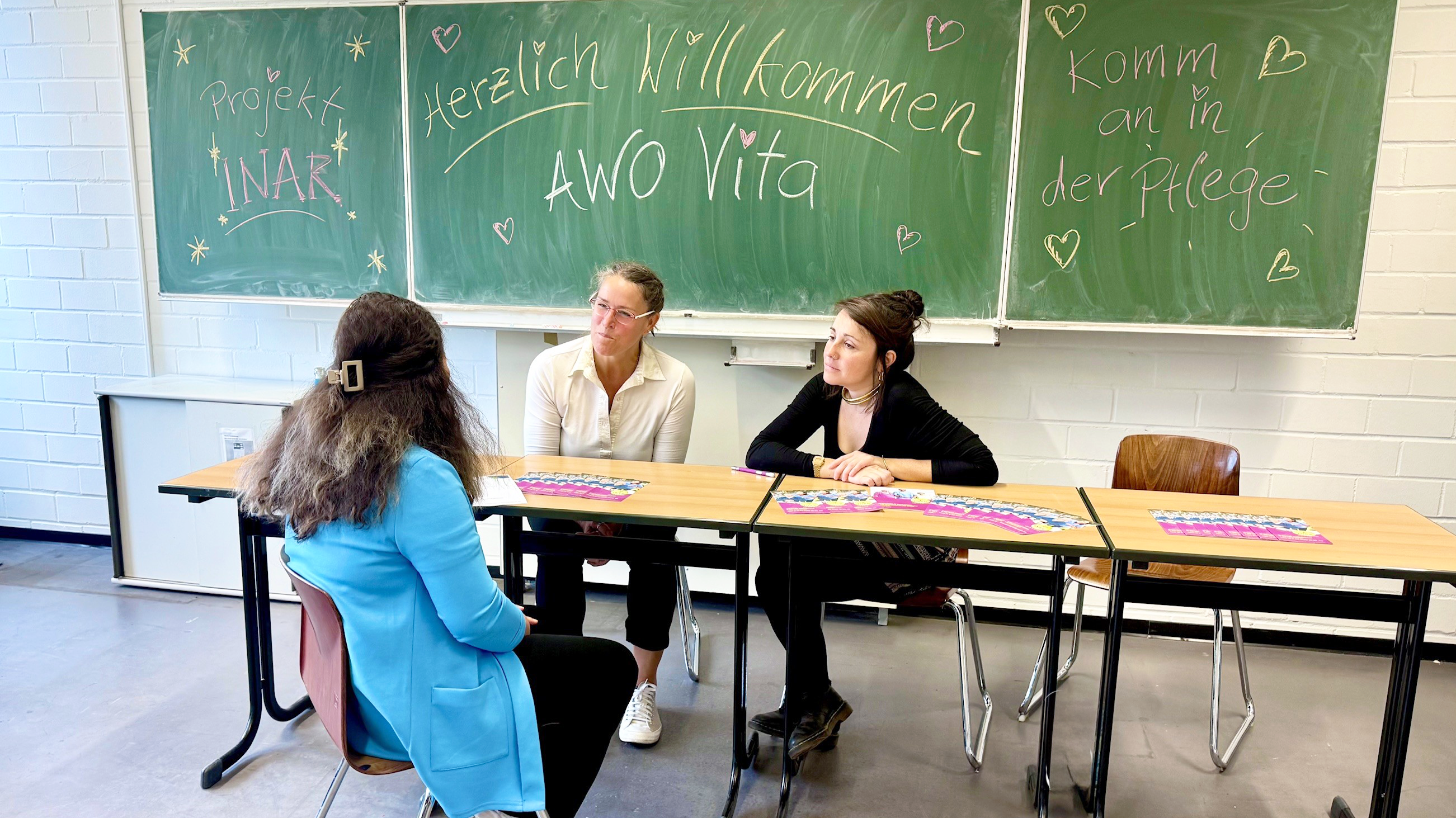  Auf dem Foto sind drei Personen an einem Tisch in einem Klassenraum zu sehen. An der grünen Tafel hinter ihnen stehen verschiedene Sätze, wie z. B. „Projekt INAR“, „Herzlich Willkommen AWO Vita“ und „Komm an in der Pflege“. Auf den Tischen liegen Informationsmaterialien.