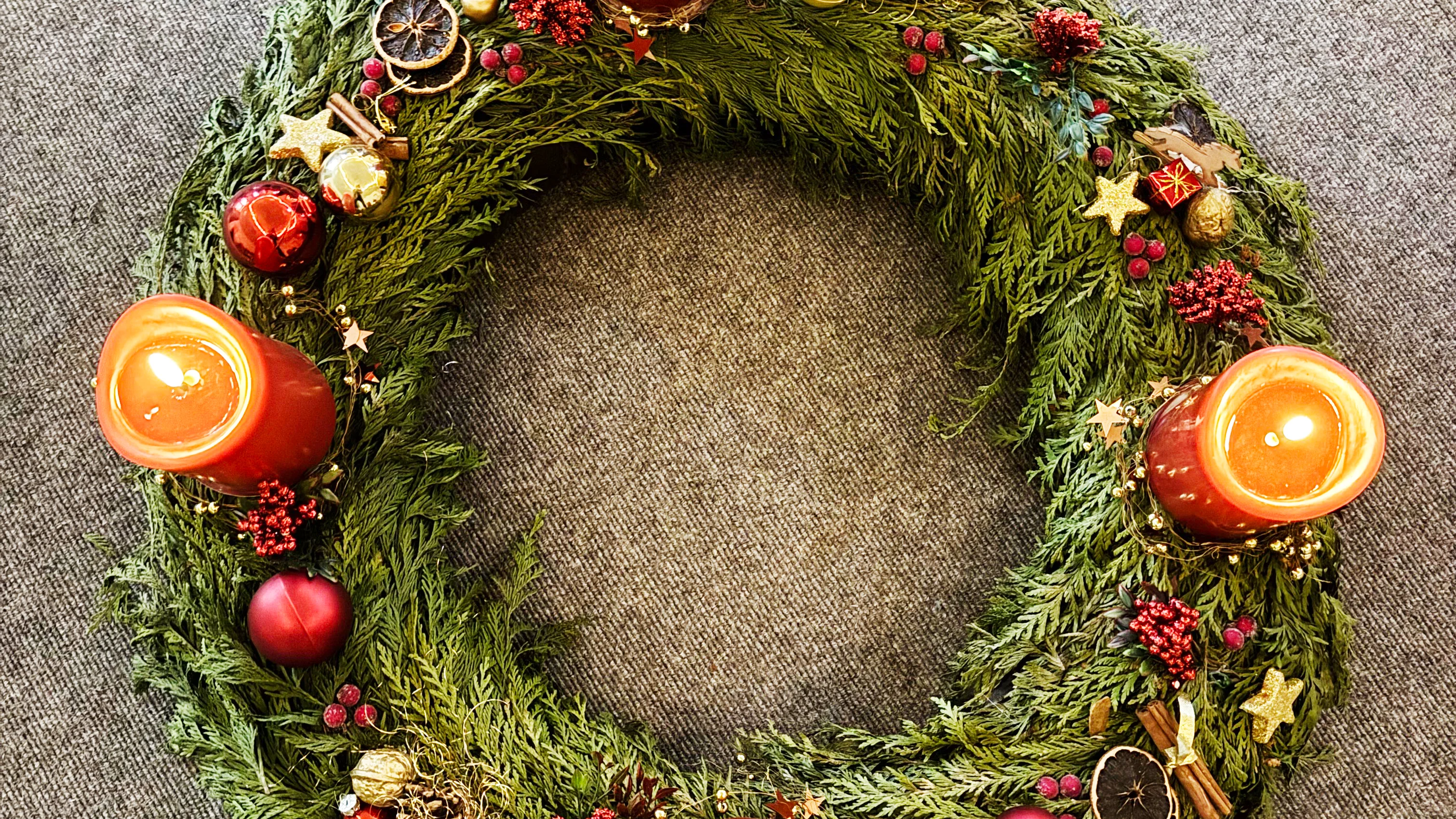 Auf dem Foto ist ein wunderschön geschmückter Adventskranz zu sehen. Der Kranz besteht aus grünen Tannenzweigen und ist mit roten und goldenen Kugeln, getrockneten Orangenscheiben, Zimtstangen, kleinen Sternen und roten Beeren dekoriert. Vier rote Kerzen sind darauf platziert, von denen drei noch nicht angezündet sind und eine bereits brennt. Der Kranz liegt auf einer grauen Oberfläche.
