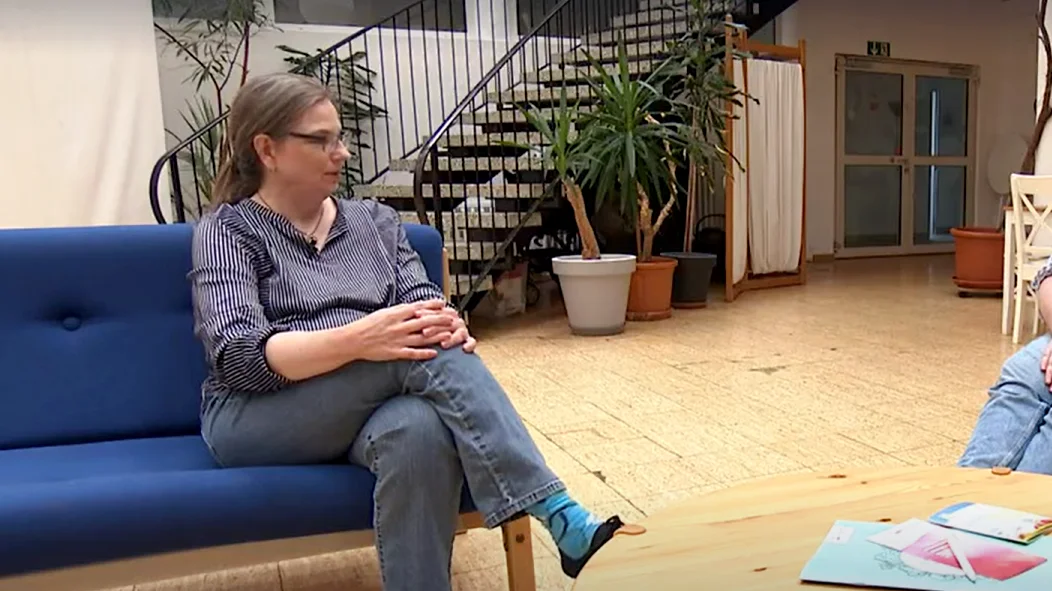 Auf dem Foto ist eine Frau zu sehen, die auf einer blauen Couch sitzt und Jeans und ein gestreiftes Hemd trägt. Sie hat eine Brille auf und die Beine übereinandergeschlagen. Neben ihr steht ein runder Holztisch, auf dem einige Gegenstände liegen. Im Hintergrund sind Pflanzen und eine Treppe zu erkennen. Es scheint sich um einen Innenraum zu handeln, möglicherweise einen Wartebereich oder eine Empfangshalle.