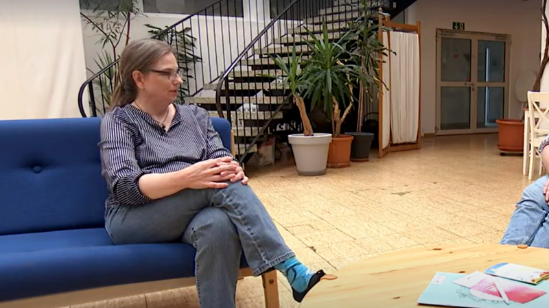 Auf dem Foto ist eine Frau zu sehen, die auf einer blauen Couch sitzt und Jeans und ein gestreiftes Hemd trägt. Sie hat eine Brille auf und die Beine übereinandergeschlagen. Neben ihr steht ein runder Holztisch, auf dem einige Gegenstände liegen. Im Hintergrund sind Pflanzen und eine Treppe zu erkennen. Es scheint sich um einen Innenraum zu handeln, möglicherweise einen Wartebereich oder eine Empfangshalle.