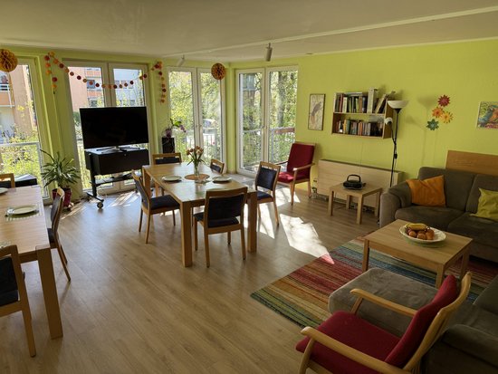 Das Bild zeigt einen hellen Raum mit großen Fenstern, durch die viel Tageslicht einfällt. Der Raum ist in warmen Farben dekoriert, hauptsächlich in Gelb- und Grüntönen. Es gibt mehrere Holztische und Stühle, die gleichmäßig im Raum verteilt sind. An der linken Wand steht ein großer Fernseher auf einem schwarzen Schrank. Rechts im Bild befindet sich eine gemütliche Sitzecke mit einem grauen Sofa, bunten Kissen und einem kleinen Tisch mit einer Obstschale darauf. Ein bunter Teppich liegt vor dem Sofa. An den Wänden hängen verschiedene Dekorationen und Regale mit Büchern. Pflanzen sind ebenfalls im Raum verteilt, was eine einladende Atmosphäre schafft.