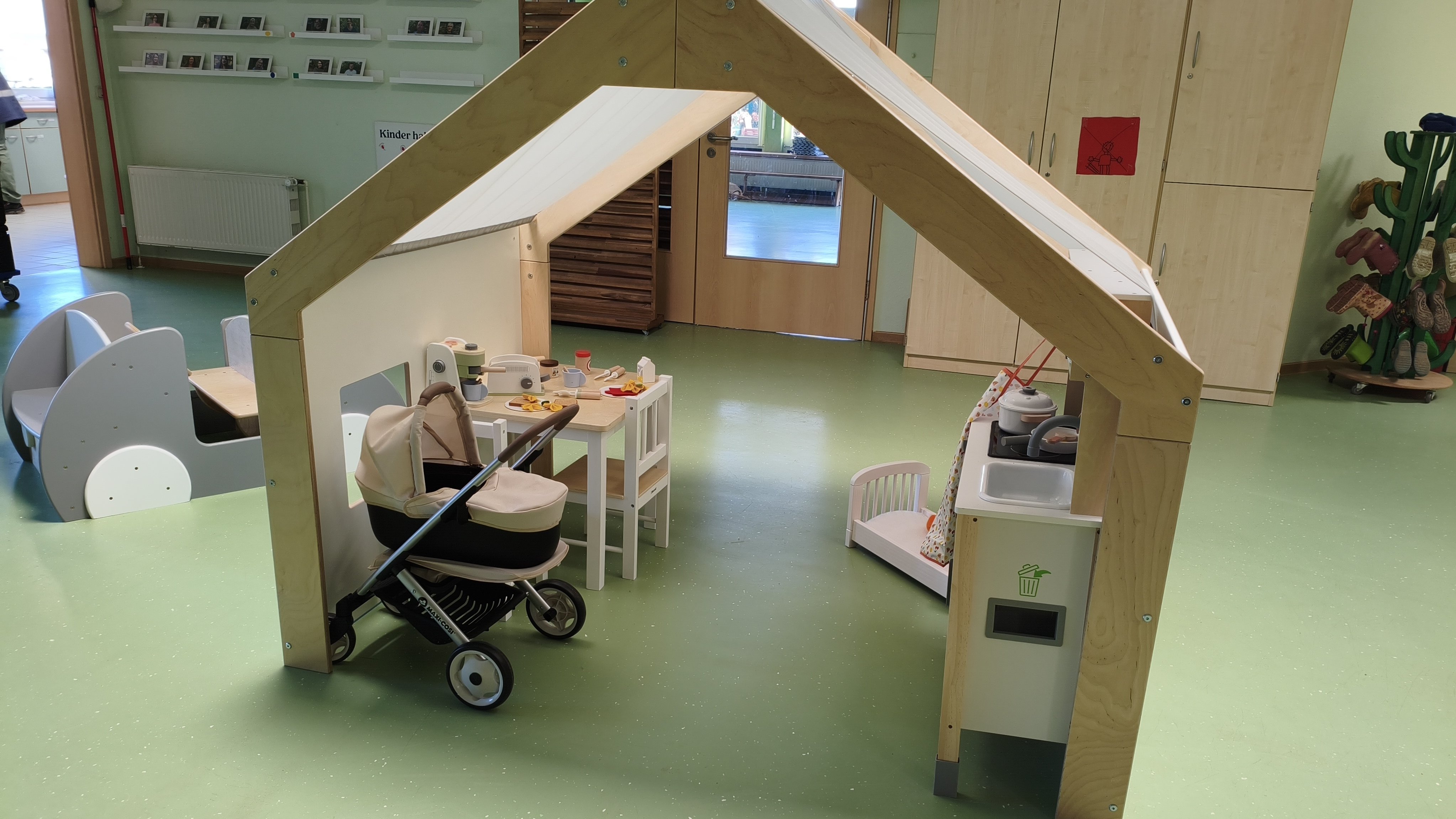 Auf dem Bild ist ein heller Spielraum für Kinder zu sehen, vermutlich in einer Kita oder einem Kindergarten. In der Mitte steht ein Holz-Spielhaus mit Dach, darunter eine kleine Spielküche, ein Kindertisch mit zwei Stühlen, Spielgeschirr und Essen sowie ein Kinderwagen.