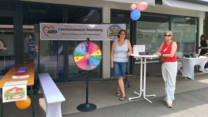 Auf dem Foto sieht man einen aufgebauten Informations- und Aktionsstand vor dem „AWO Familienzentrum Baumberg – Kitas Kunterbunt & Villa Regenbogen“. Links steht eine Bierzeltgarnitur mit Bank, Luftballon und einem angeklebten Kinderbild, in der Mitte ein buntes Glücksrad, rechts ein Stehtisch mit Infomaterial sowie im Hintergrund einen weiteren Tisch mit Utensilien