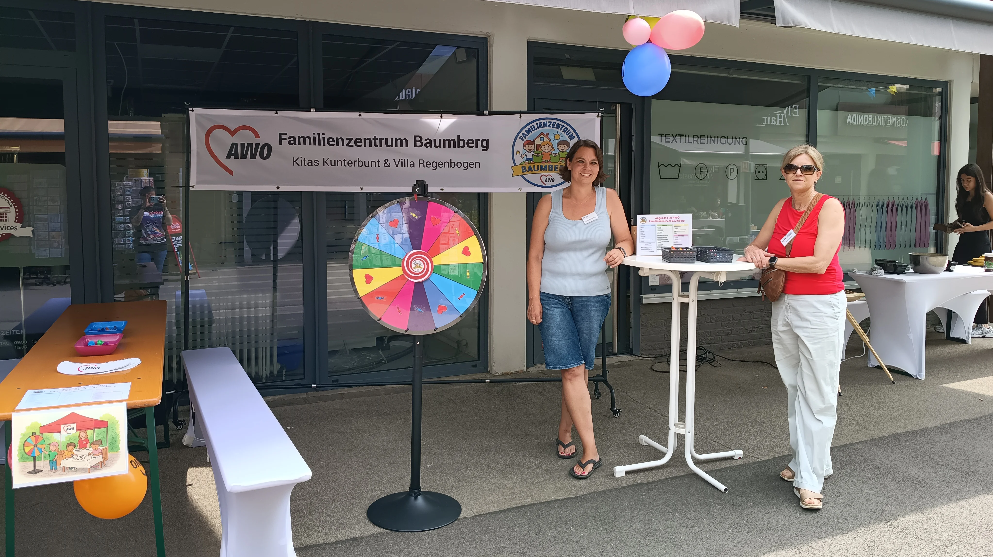 Auf dem Foto sieht man einen aufgebauten Informations- und Aktionsstand vor dem „AWO Familienzentrum Baumberg – Kitas Kunterbunt & Villa Regenbogen“. Links steht eine Bierzeltgarnitur mit Bank, Luftballon und einem angeklebten Kinderbild, in der Mitte ein buntes Glücksrad, rechts ein Stehtisch mit Infomaterial sowie im Hintergrund einen weiteren Tisch mit Utensilien