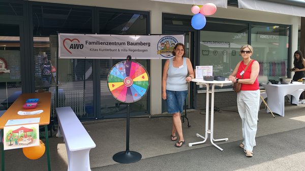 Auf dem Foto sieht man einen aufgebauten Informations- und Aktionsstand vor dem „AWO Familienzentrum Baumberg – Kitas Kunterbunt & Villa Regenbogen“. Links steht eine Bierzeltgarnitur mit Bank, Luftballon und einem angeklebten Kinderbild, in der Mitte ein buntes Glücksrad, rechts ein Stehtisch mit Infomaterial sowie im Hintergrund einen weiteren Tisch mit Utensilien