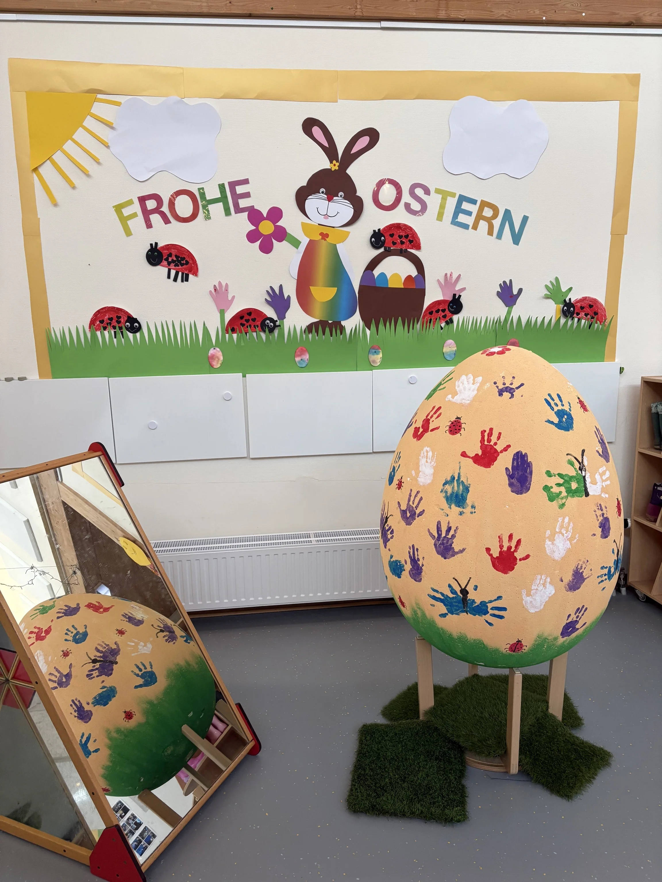 Auf dem Foto sieht man eine österliche Dekoration in einer Kita oder ähnlichen Einrichtung. Im Hintergrund hängt ein großes Wandbild mit der Aufschrift „Frohe Ostern“, einem Osterhasen, Marienkäfern und buntem Gras. Im Vordergrund steht ein großes, gelb bemaltes Osterei auf einem Holzständer, das mit vielen bunten Handabdrücken von Kindern verziert ist. Links daneben steht ein Spiegel, in dem sich das Osterei noch einmal spiegelt.