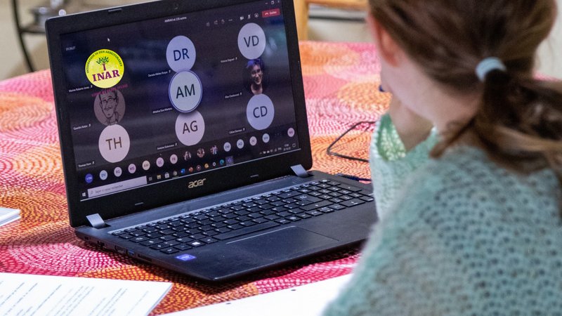 Zu sehen ist eine Person von hinten, die an einem Tisch vor einem Acer-Laptop sitzt und an einer Online-Videokonferenz teilnimmt. Auf dem Bildschirm sind mehrere Teilnehmende als Initialen beziehungsweise Profilbilder zu sehen; außerdem ist das Logo des AWO Projekts INAR eingeblendet.