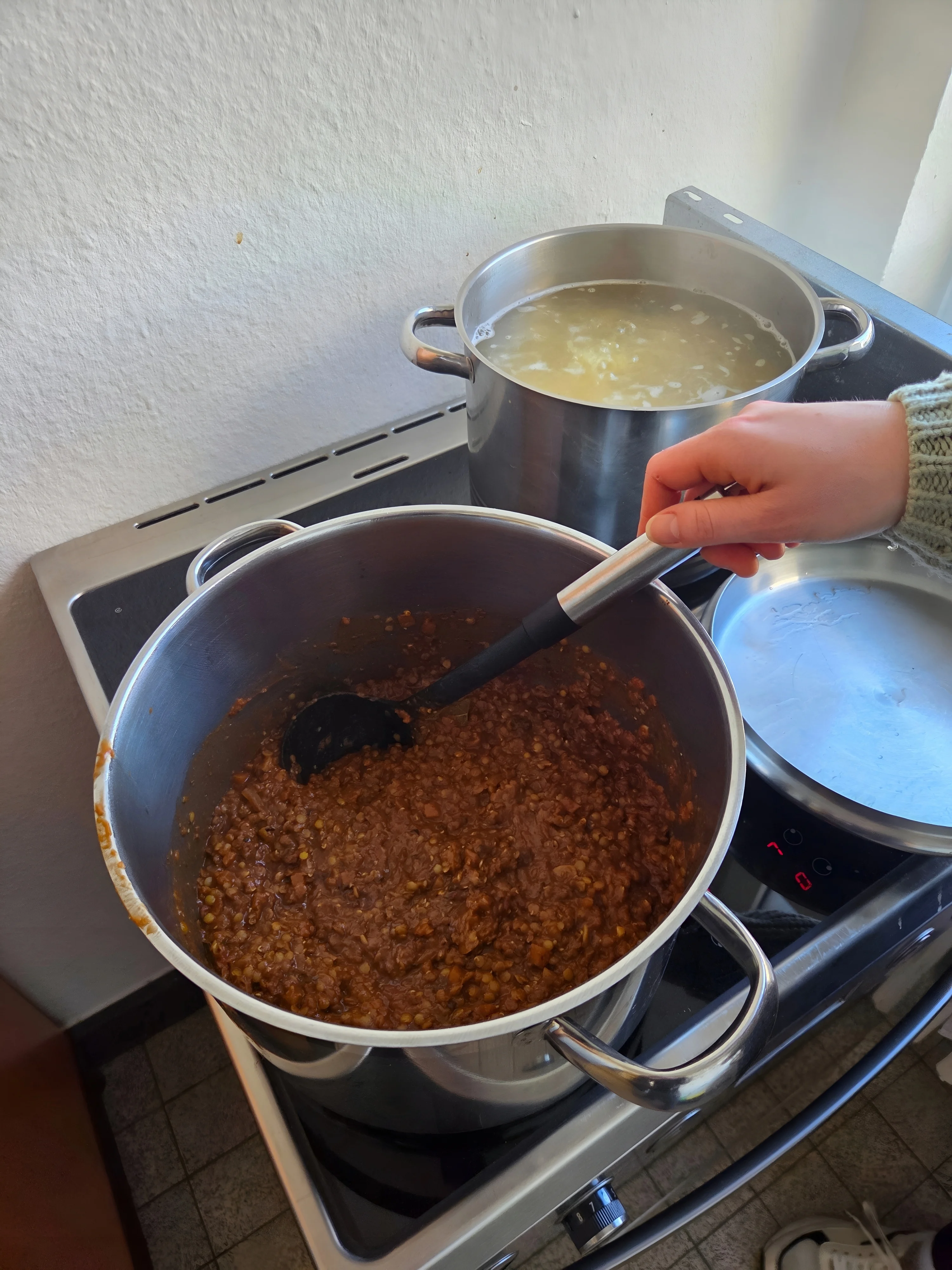 Auf dem Bild sieht man zwei große Kochtöpfe auf einem Herd: In dem vorderen Topf wird eine dicke, rote Linsen‑ oder Bolognese‑Artige Soße mit einer Kelle umgerührt, im hinteren Topf kochen Nudeln oder Kartoffeln in Wasser.