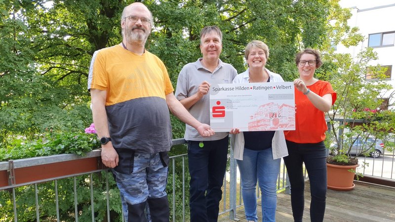 In dem Bild sind vier Personen auf einem Balkon zu sehen, im Hintergrund ist viel Grün. Sie halten einen Scheck von der “Sparkasse Hilden Ratingen Velbert” in den Händen.