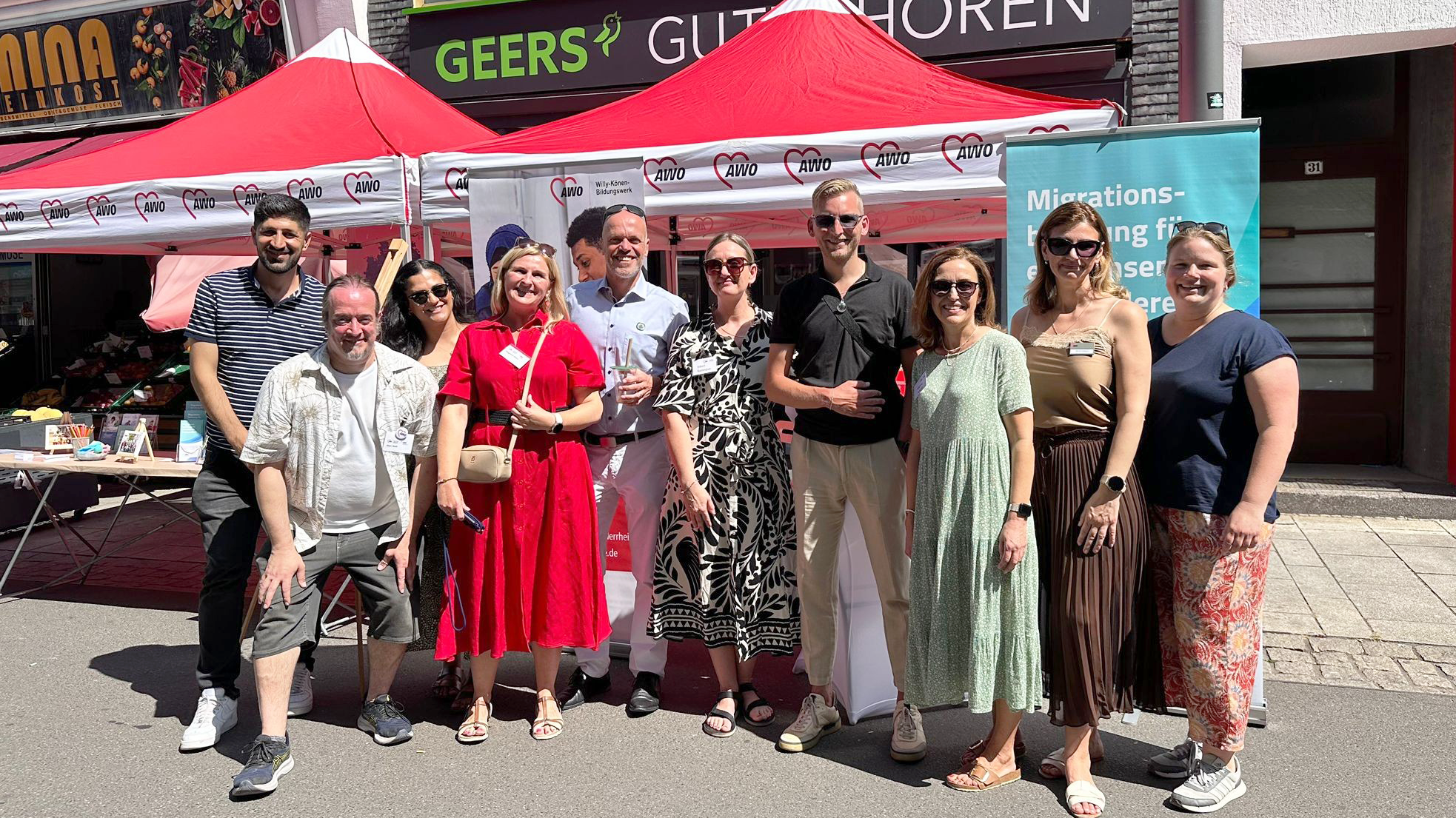 Auf dem Foto ist eine Gruppe von Menschen zu sehen, die gemeinsam vor einem Info-Stand der AWO (Arbeiterwohlfahrt) bei einer öffentlichen Veranstaltung stehen – vermutlich ein Straßen- oder Kulturfest. Der Stand ist mit roten Pavillons und AWO-Logos geschmückt. Ein Banner im Hintergrund weist auf das Thema „Migrationsberatung“ hin. Die Gruppe wirkt fröhlich und entspannt, viele lächeln in die Kamera. Es ist sonnig, die Menschen tragen sommerliche Kleidung. Im Hintergrund sind Geschäfte wie „GEERS GUT HÖREN“ und „MINA Feinkost“ zu erkennen. Das Bild vermittelt eine offene und gemeinschaftliche Atmosphäre. Wahrscheinlich handelt es sich um das „Fest der Kulturen“ in Grevenbroich, wie der Dateiname vermuten lässt.