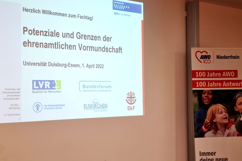  Auf dem Bild ist eine Präsentationsfolie zu sehen, die auf eine Leinwand projiziert wird, sowie ein Banner im rechten Teil des Bildes. ?️ Präsentationsfolie Die Folie ist in deutscher Sprache und trägt die Überschrift: "Herzlich Willkommen zum Fachtag!" "Potenziale und Grenzen der ehrenamtlichen Vormundschaft" Darunter steht der Veranstaltungsort und das Datum: "Universität Duisburg-Essen, 1. April 2022" Im unteren Bereich der Folie sind Logos verschiedener Organisationen oder Projekte abgebildet, darunter: LVR Bundesforum Vormundschaft und Pflegeelternschaft Der Kinderschutzbund Diözesanverband Aachen Ein Logo mit der Aufschrift Kreis EUSKIRCHEN SKF Am unteren linken Rand sind noch Textteile zu erkennen wie "Forschungs- und Entwicklungsprojekt" und "Dr. Michael Maier". ? Banner Rechts neben der Projektionsfläche ist ein Teil eines großen Banners sichtbar, auf dem steht: AWO (Logo) Niederhein 100 Jahre AWO 100 Jahre Antwor[t] (der Text ist teilweise abgeschnitten). Unten auf dem Banner sind undeutliche Gesichter von Personen, möglicherweise Kindern, zu sehen. Zusammenfassend handelt es sich um eine Aufnahme von einem Fachtag oder einer Veranstaltung zum Thema ehrenamtliche Vormundschaft an der Universität Duisburg-Essen, unterstützt unter anderem von der AWO.