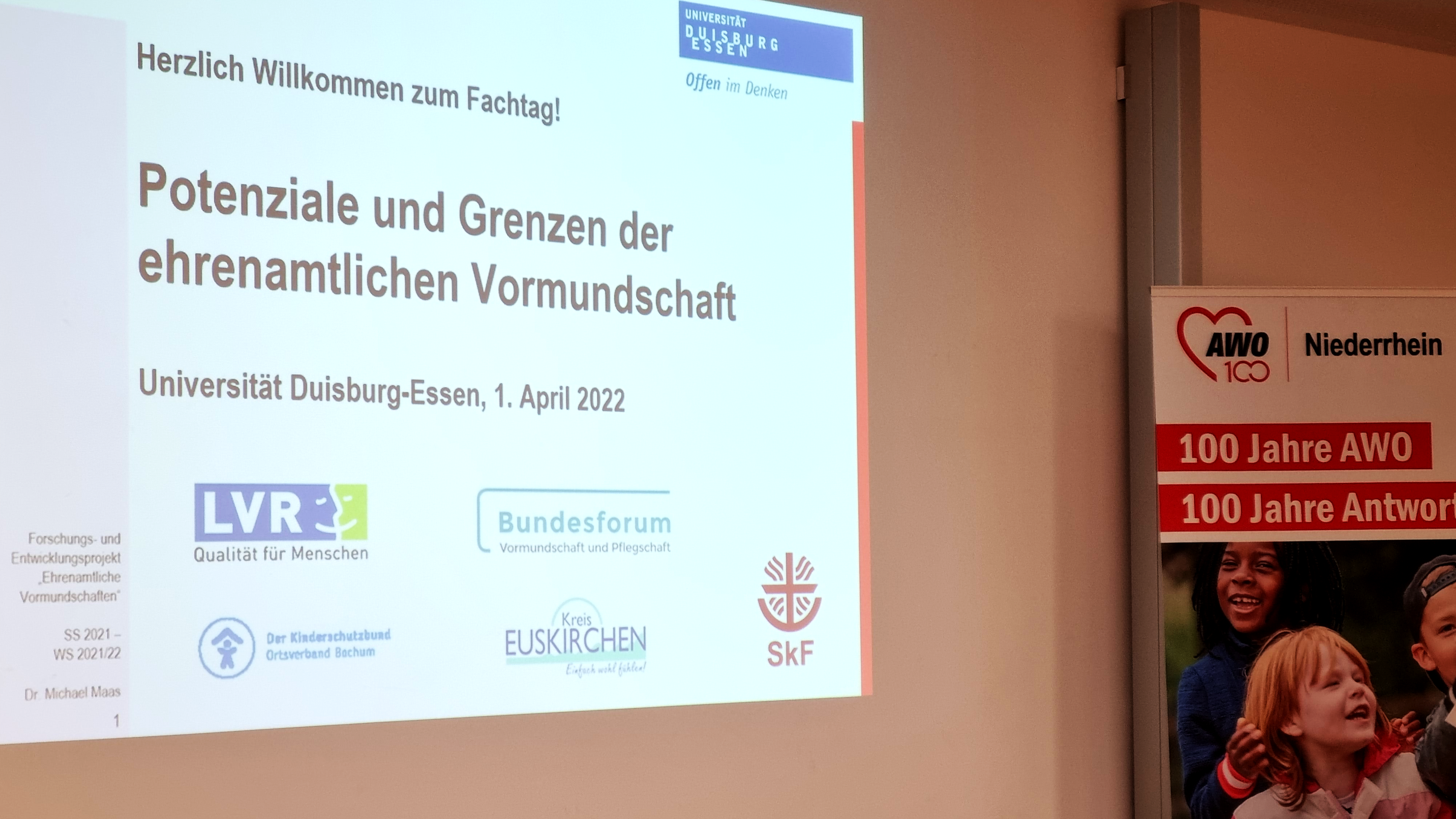  Auf dem Bild ist eine Präsentationsfolie zu sehen, die auf eine Leinwand projiziert wird, sowie ein Banner im rechten Teil des Bildes. ?️ Präsentationsfolie Die Folie ist in deutscher Sprache und trägt die Überschrift: "Herzlich Willkommen zum Fachtag!" "Potenziale und Grenzen der ehrenamtlichen Vormundschaft" Darunter steht der Veranstaltungsort und das Datum: "Universität Duisburg-Essen, 1. April 2022" Im unteren Bereich der Folie sind Logos verschiedener Organisationen oder Projekte abgebildet, darunter: LVR Bundesforum Vormundschaft und Pflegeelternschaft Der Kinderschutzbund Diözesanverband Aachen Ein Logo mit der Aufschrift Kreis EUSKIRCHEN SKF Am unteren linken Rand sind noch Textteile zu erkennen wie "Forschungs- und Entwicklungsprojekt" und "Dr. Michael Maier". ? Banner Rechts neben der Projektionsfläche ist ein Teil eines großen Banners sichtbar, auf dem steht: AWO (Logo) Niederhein 100 Jahre AWO 100 Jahre Antwor[t] (der Text ist teilweise abgeschnitten). Unten auf dem Banner sind undeutliche Gesichter von Personen, möglicherweise Kindern, zu sehen. Zusammenfassend handelt es sich um eine Aufnahme von einem Fachtag oder einer Veranstaltung zum Thema ehrenamtliche Vormundschaft an der Universität Duisburg-Essen, unterstützt unter anderem von der AWO.