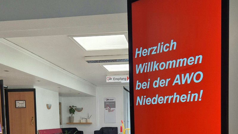 Auf dem Foto ist eine digitale Infostele in einer Geschäftsstelle der AWO Niederrhein zu sehen. Die Stele hat einen großen, hochformatigen Bildschirm mit rotem Hintergrund und weißer Schrift. Der angezeigte Text lautet: „Herzlich Willkommen bei der AWO Niederrhein!“ Unten auf der Stele ist das AWO-Logo mit dem Schriftzug „AWO Niederrhein“ zu sehen. Im Hintergrund erkennt man den Empfangsbereich mit einem Wartebereich, Pflanzen und einigen Wegweisern. Die Umgebung wirkt freundlich und einladend.